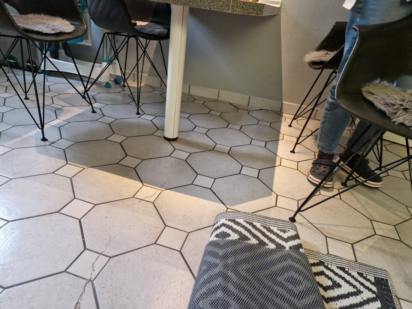 Sechseckige graue Bodenfliesen; Stühle um einen Tisch, rechts sitzt eine Person in Jeans und Crocs.