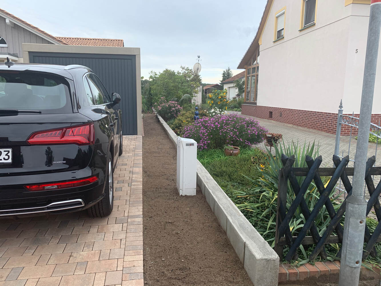 Schwarzer SUV steht auf gepflasterter Einfahrt, daneben Garten mit bunten Blumen vor Haus.