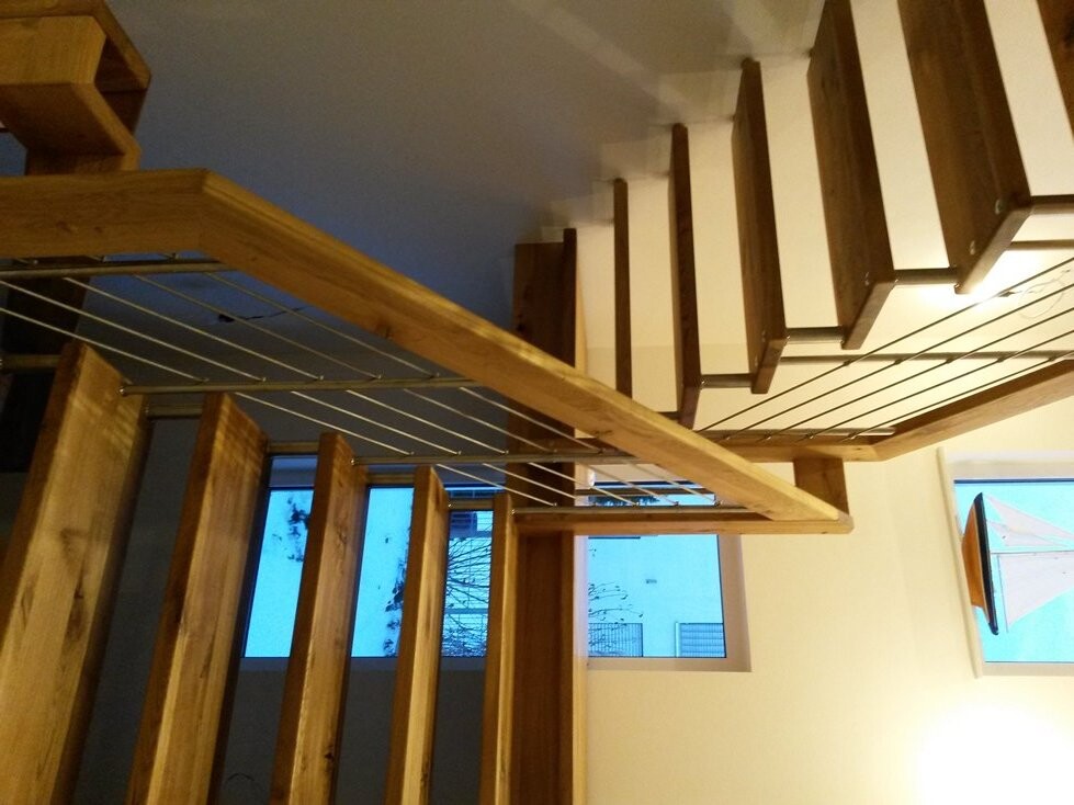 Moderne Holztreppe mit Geländer aus Metallseilen im Innenraum, Fenster sichtbar.