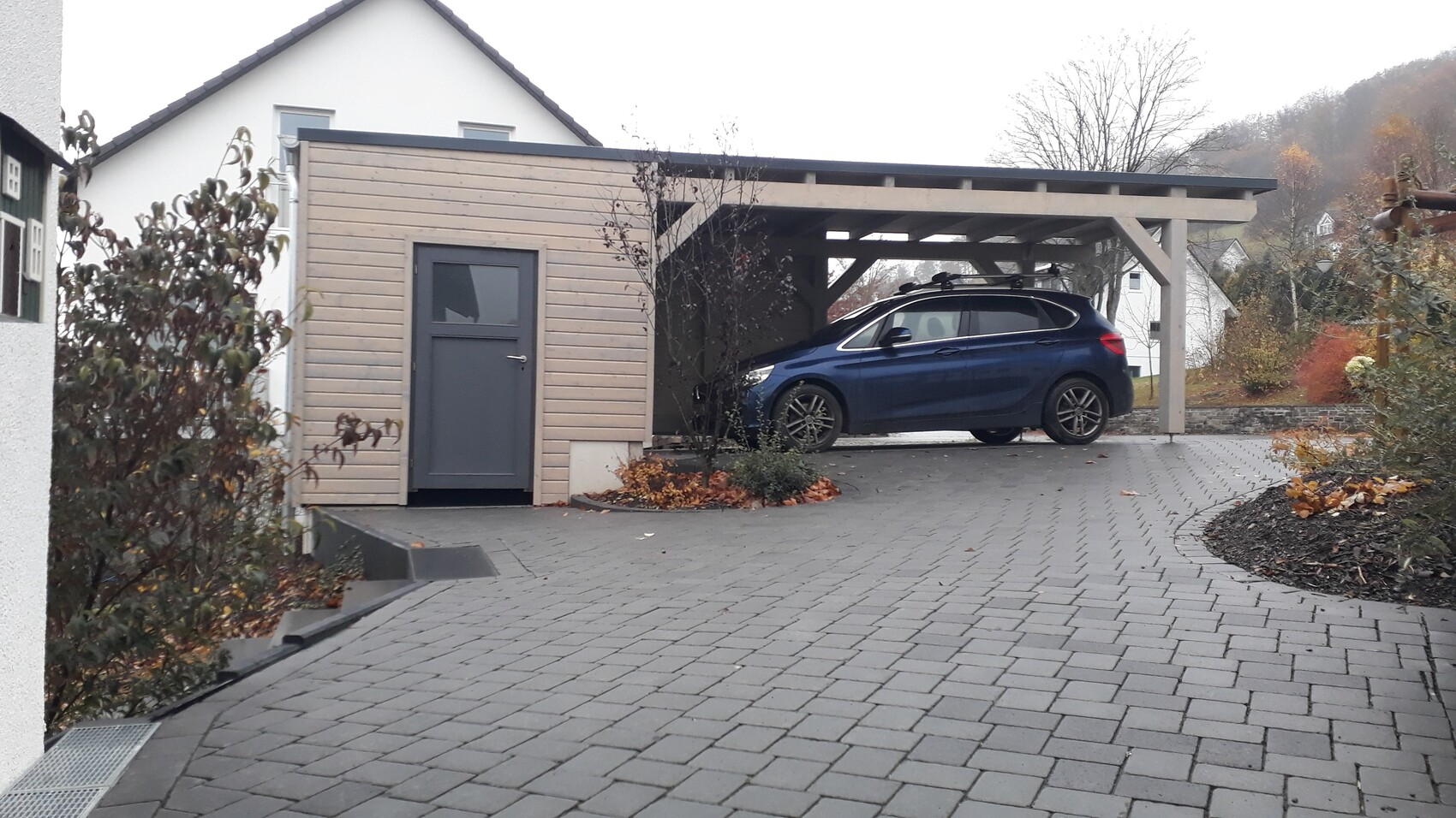 Blauer Wagen unter Carport aus Holzbohlen neben Haus, gepflasterte Einfahrt.