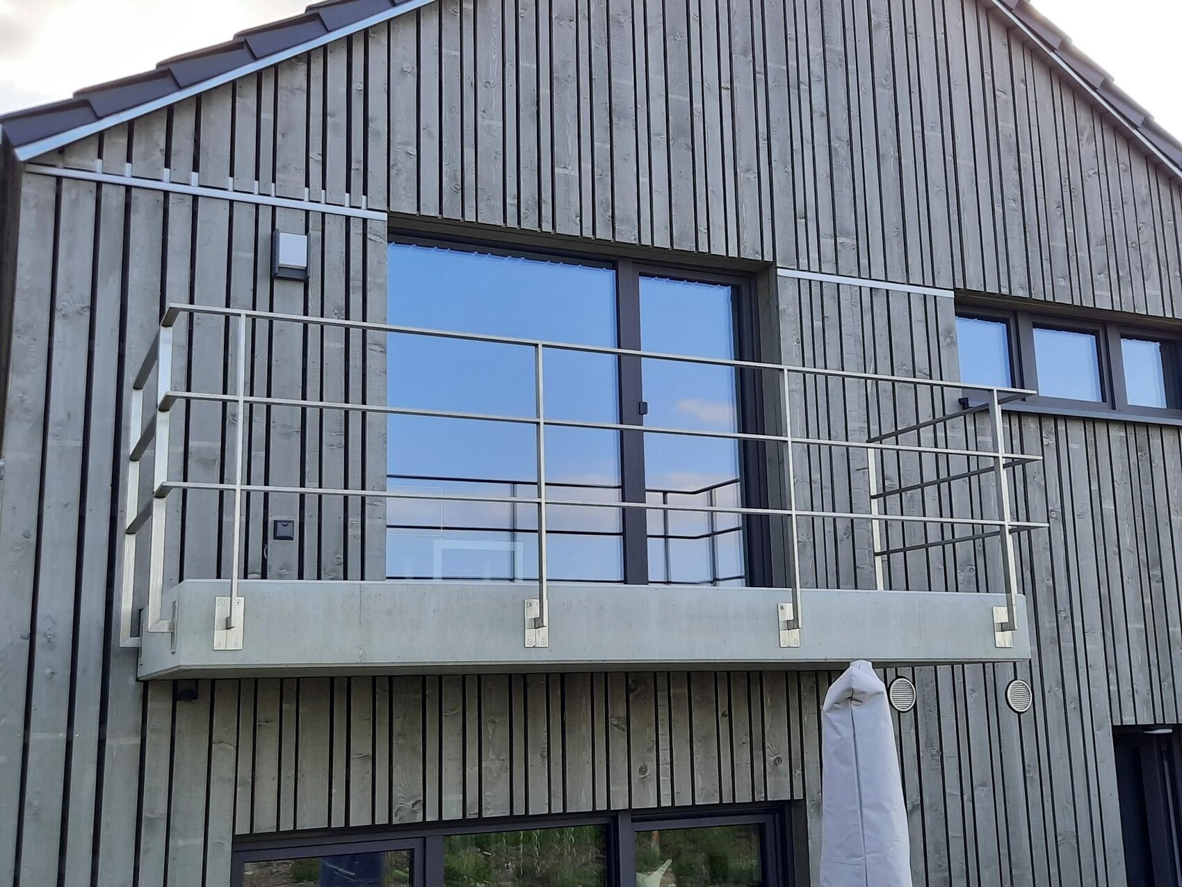 Moderne Fassade aus vertikalen Holzlatten; Balkon mit Betonboden und Metallgeländer vor Glasfenstern.