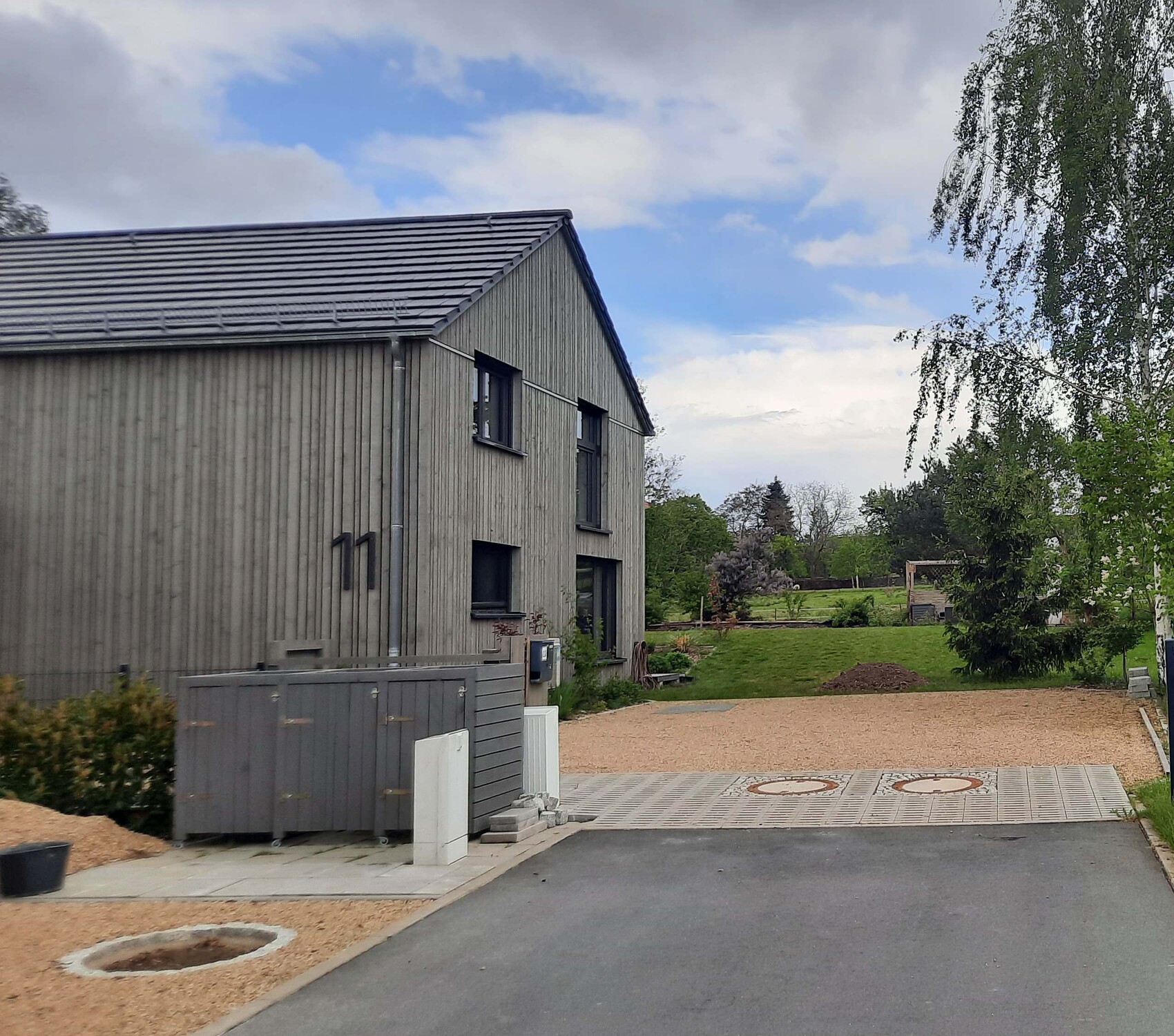 Zweistöckiges graues Holzhaus am Hof; Parkplatzfläche, Kieswege, Garten und Bäume im Hintergrund.