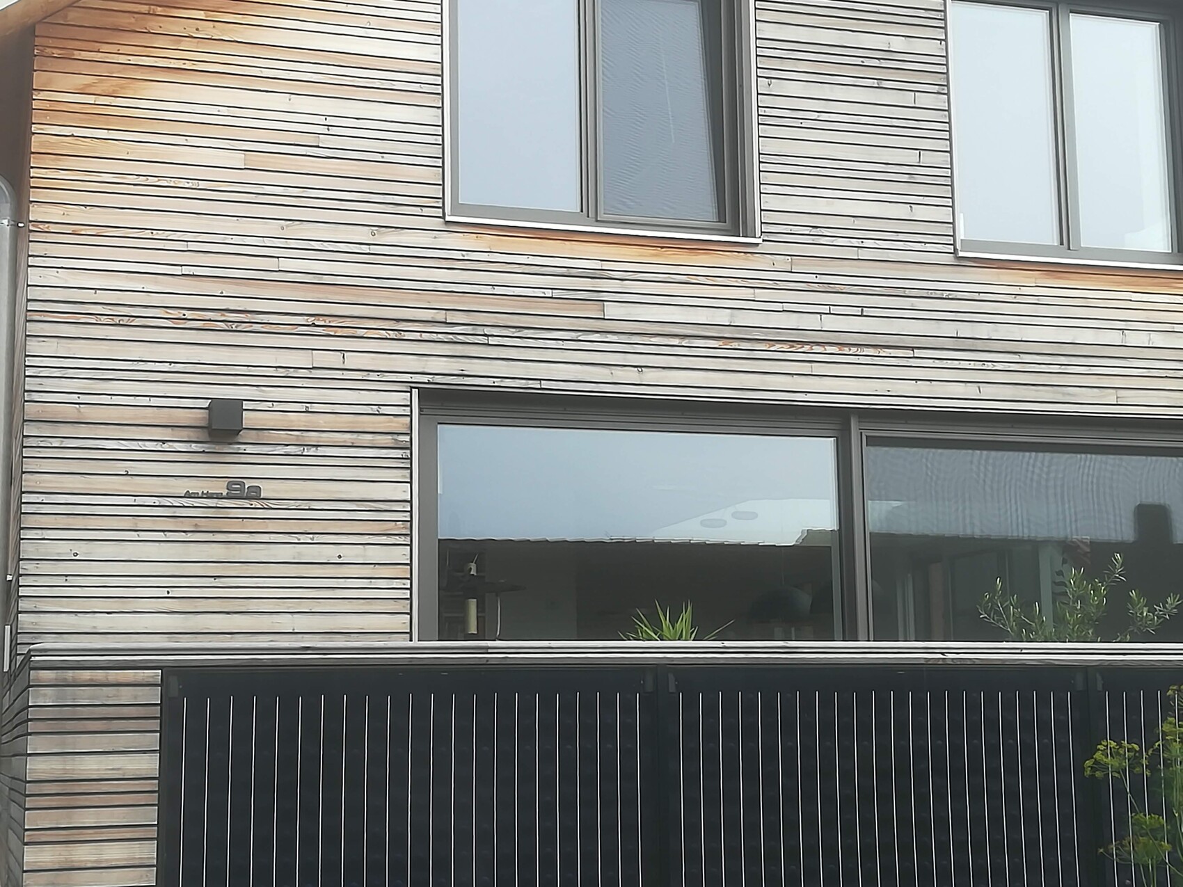 Aussenfassade aus horizontalen Holzlatten, grosse Fenster, schwarzer Metallzaun unten