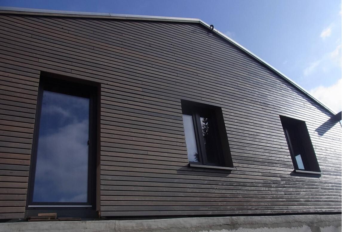 Außenansicht eines Hauses mit brauner Holzverkleidung, Tür links, zwei Fenster rechts.