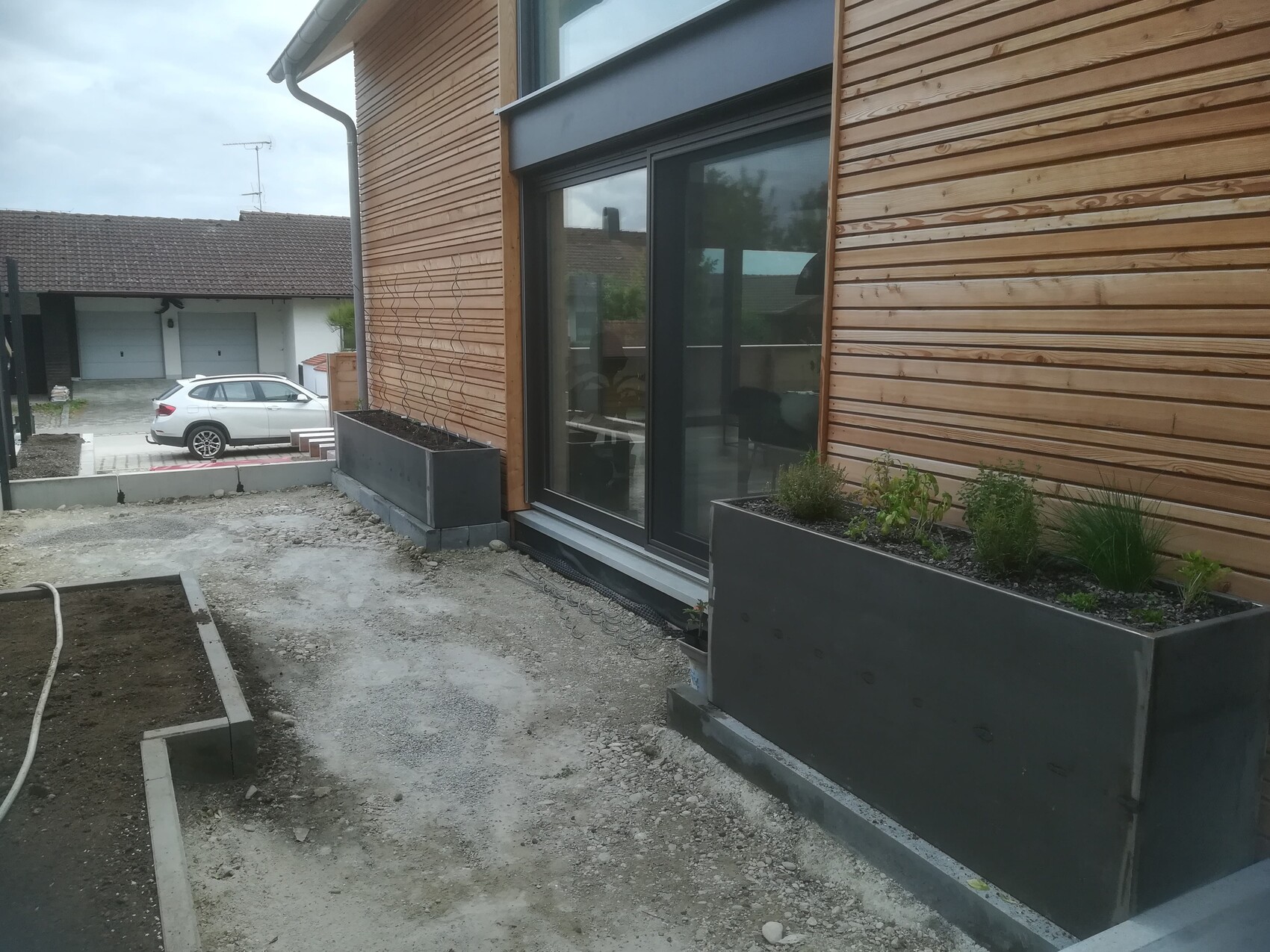Moderne Holzfassade mit großen Glasfenstern und rechteckigen Metallpflanzkästen mit Pflanzen.
