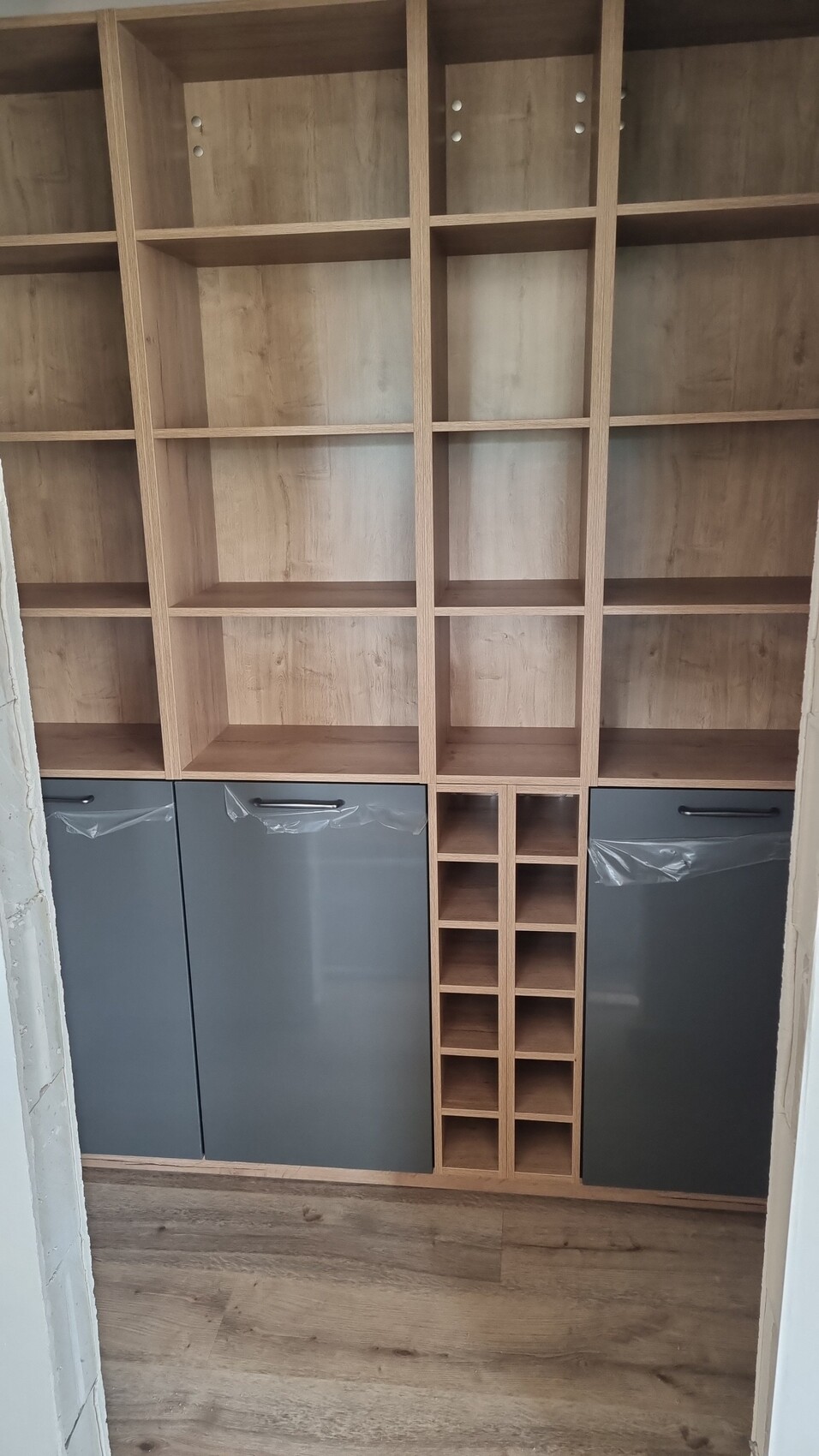 Großes Holzregal mit vielen offenen Fächern, mittig kleiner Flaschenhalter, graue Unterschränke.