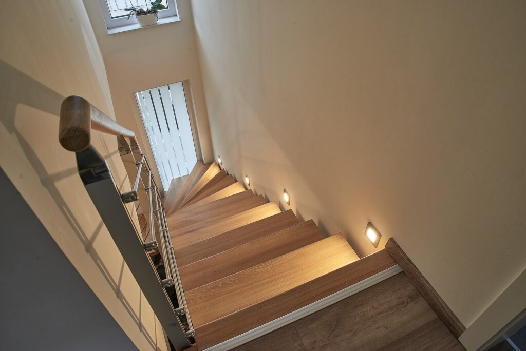Helle Holztreppe im Treppenhaus mit Metallgeländer und Wandlichtspots.