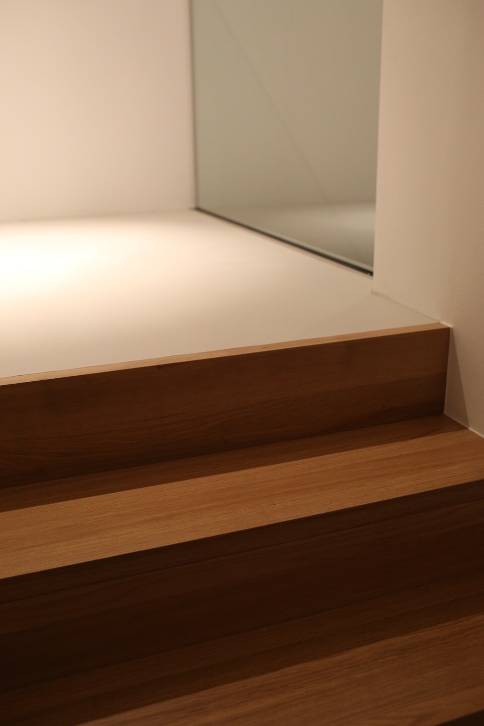 Dreistufige Holztreppe in einem hellen Raum mit weißen Wänden und einer Glasfront am oberen Rand.