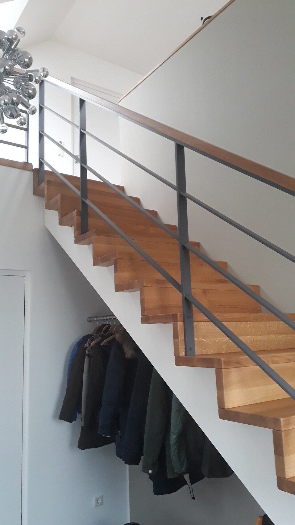 Innenansicht: Treppe mit Holzstufen, grauem Geländer und Mantelgarderobe darunter.