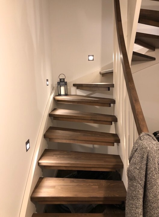 Holztreppe mit offenen Stufen in heller Nische, Wandlichter, Laterne und Mantel am Geländer.