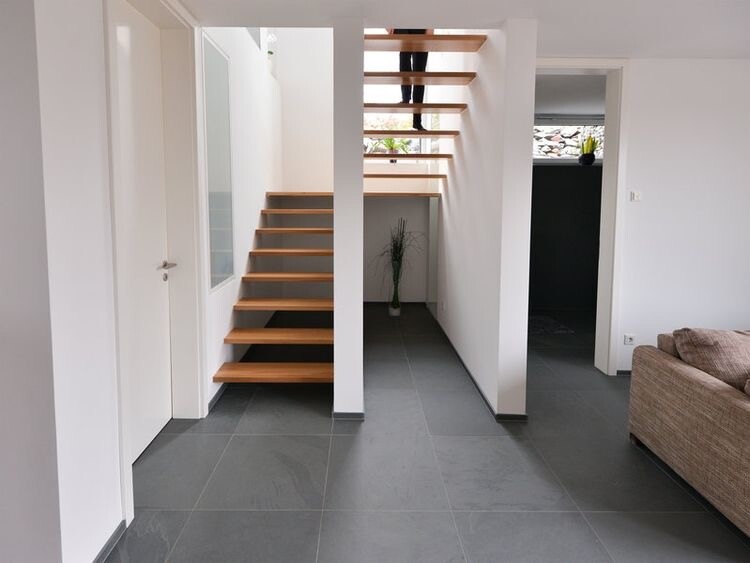Moderne Eingangshalle mit offener Holztreppe, grauen Fliesen und Couch rechts.
