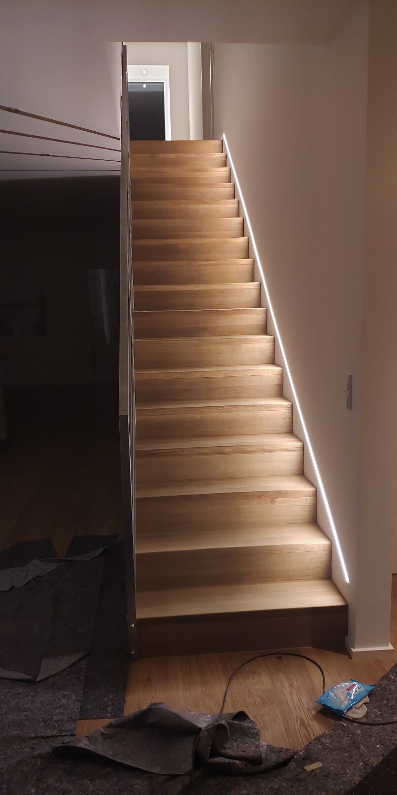Holztreppe nach oben, rechte Wand mit LED-Lichtlinie, linkes Geländer, dunkler Raum links.