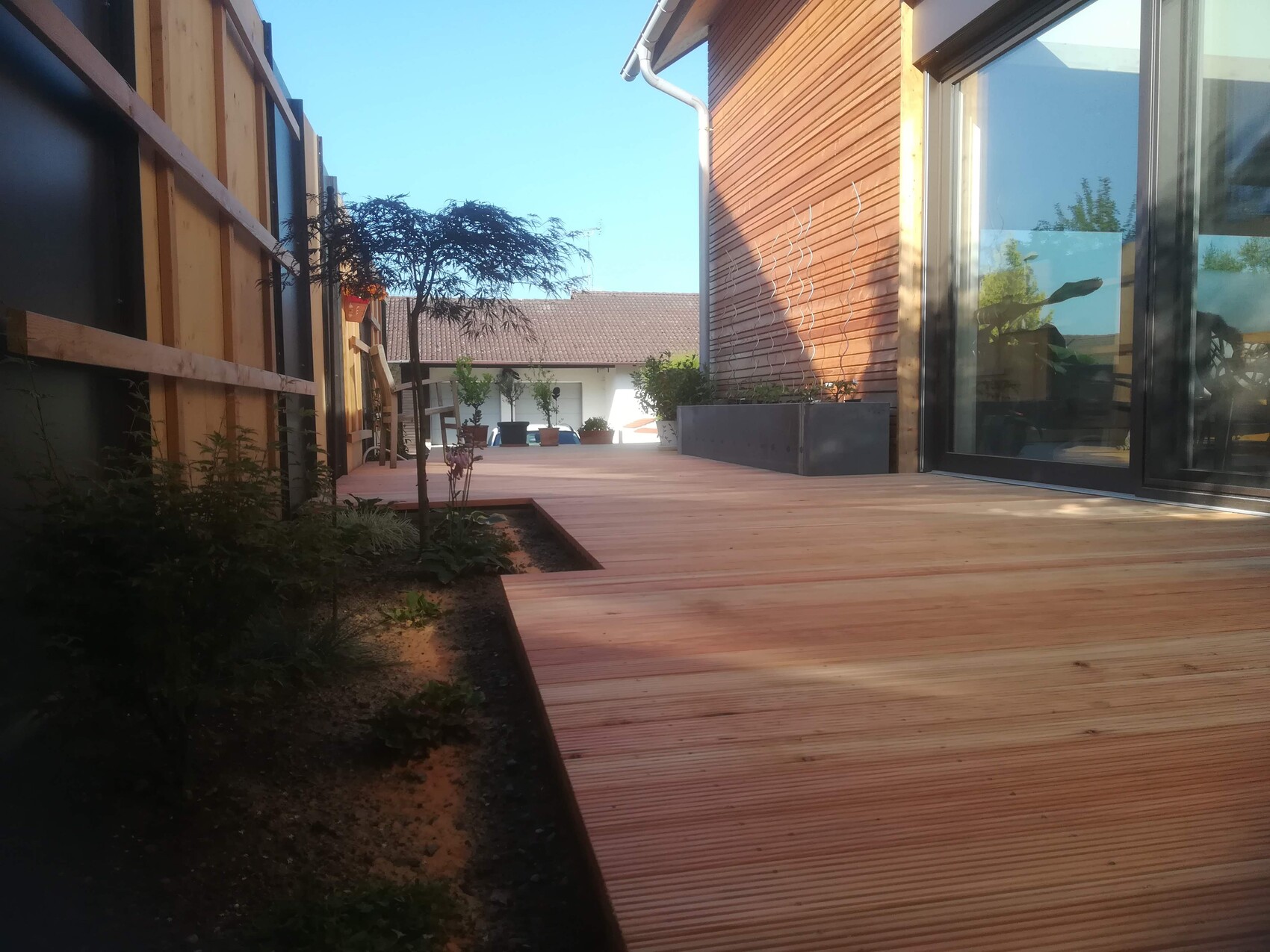 Holzdeck aus Planken zwischen Gebäuden, rechts verglaste Türen, Pflanzen und Baum im Hintergrund.