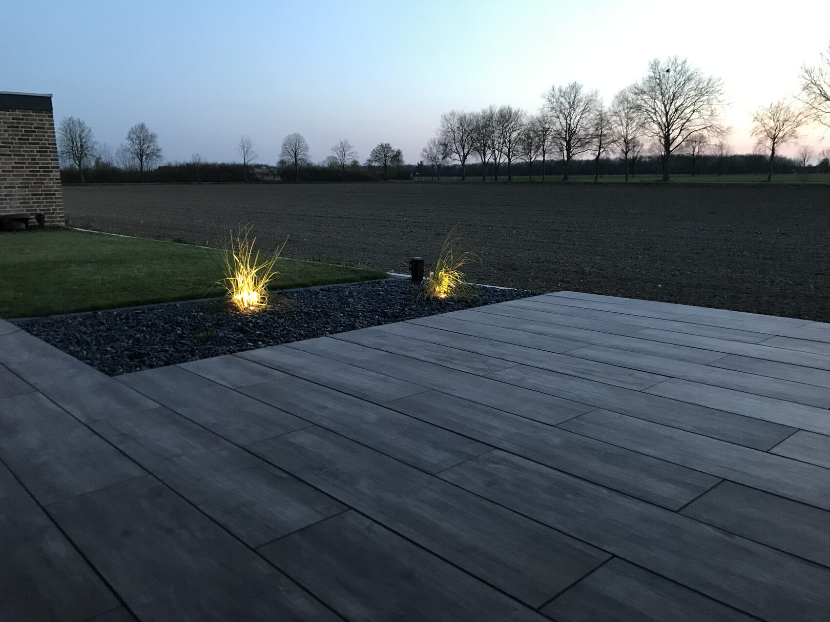 Holzterrasse mit beleuchteten Ziergräsern in Kiesbett, Blick aufs Feld und Bäume bei Dämmerung.