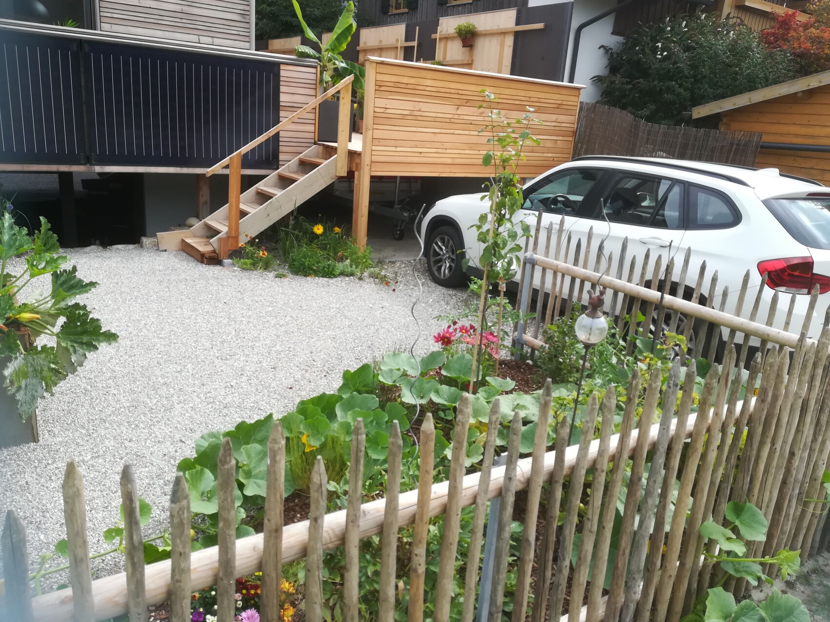 Vorgarten mit Holzdeck, Treppe, Kiesweg, Beetpflanzen und weißem SUV am Rand.