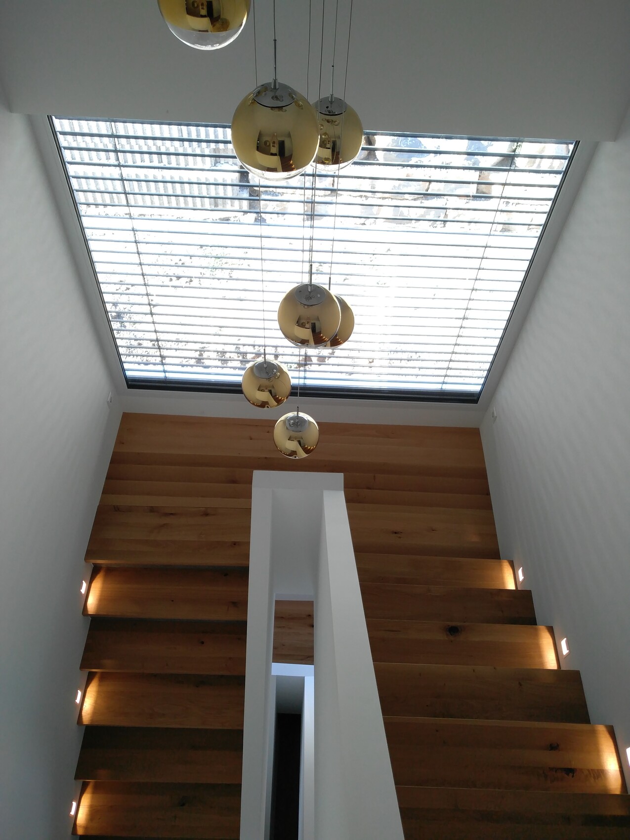 Blick von oben auf moderne Treppe mit Holzstufen, weißer Stütze und goldenen Pendelleuchten.