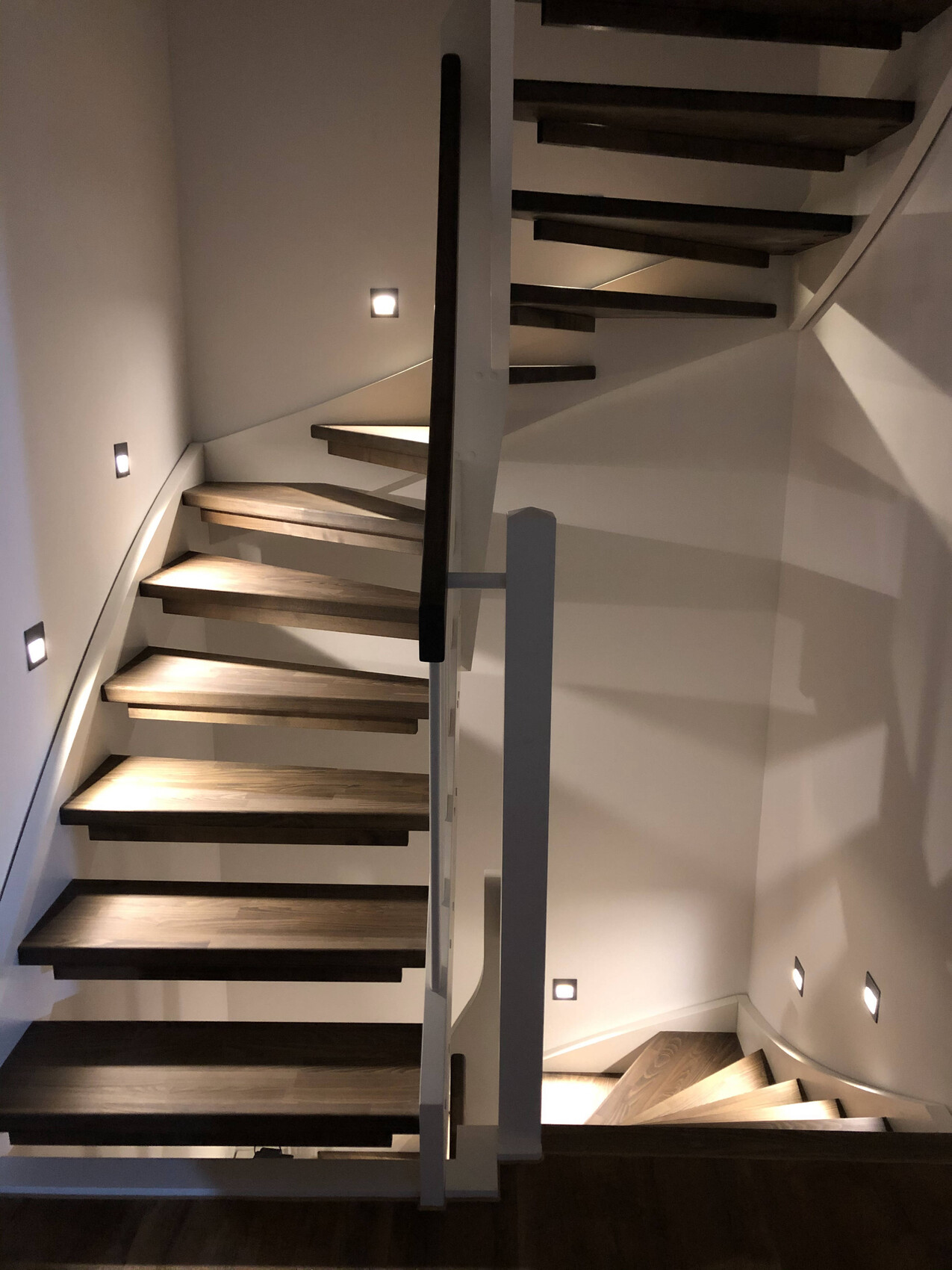 Moderne Holztreppe mit offenen Stufen, weißem Geländer und Wand-LED-Lampen im Innenraum.