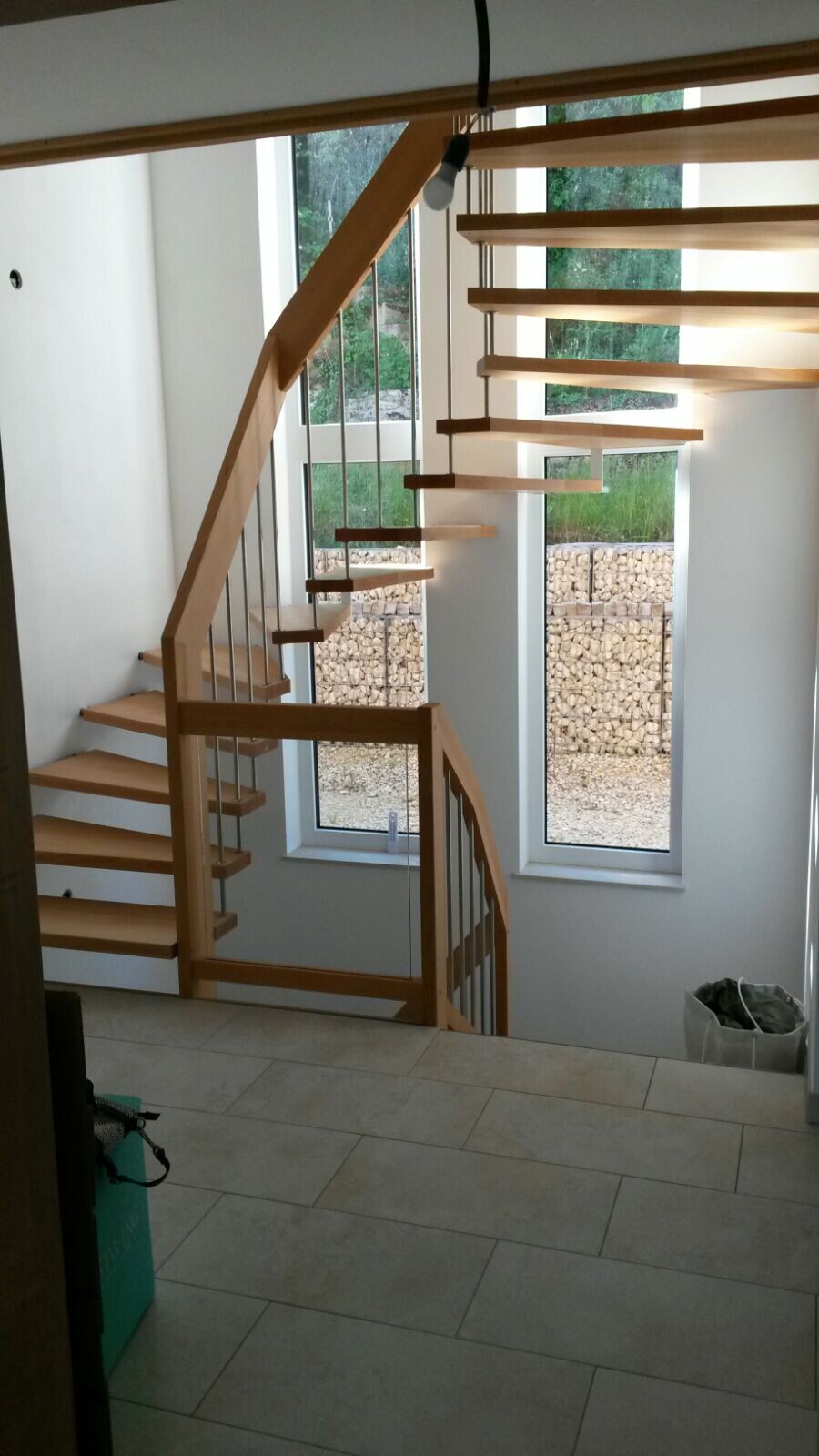 Moderne Holztreppe mit offenen Stufen und Metall-Geländer vor großen Fenstern; Außenbereich sichtbar.