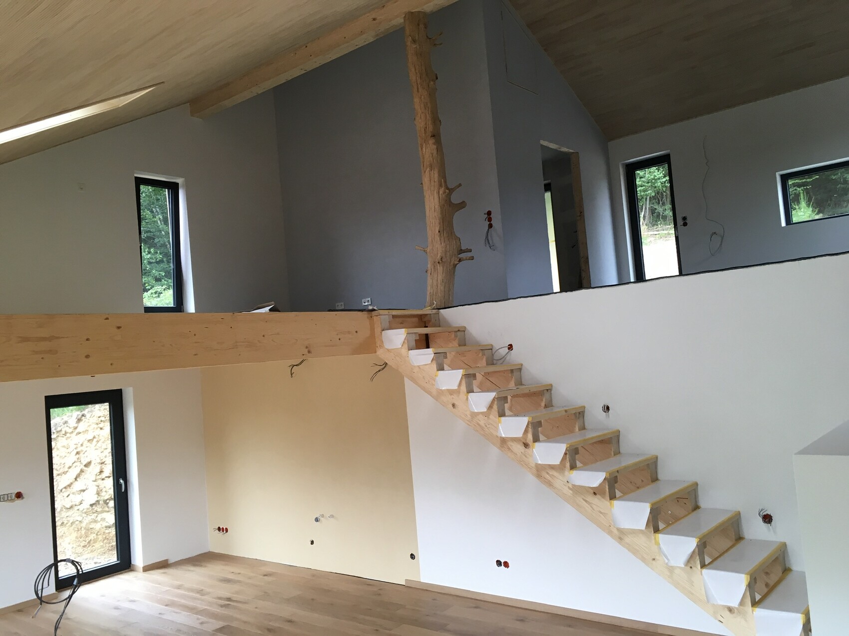 Offener Innenraum im Bau mit schwebender Holztreppe, Baumstamm-Säule und großen Fenstern.