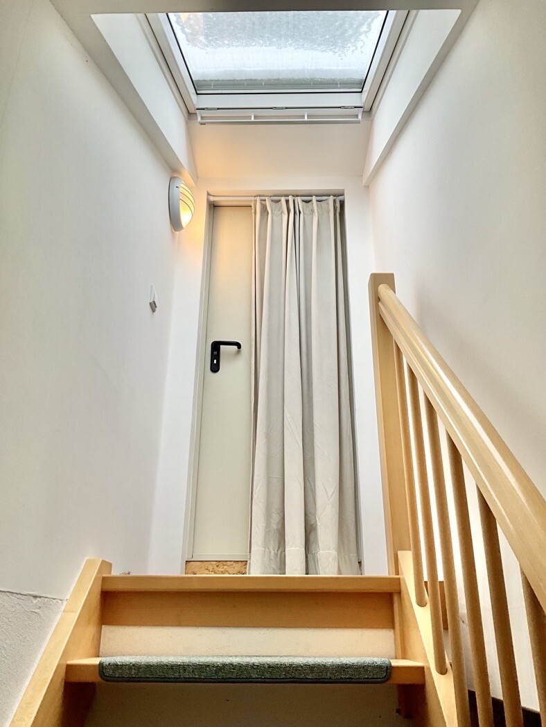 Stufen mit Holzgeländer, Tür hinter grauem Vorhang; Dachfenster oben.