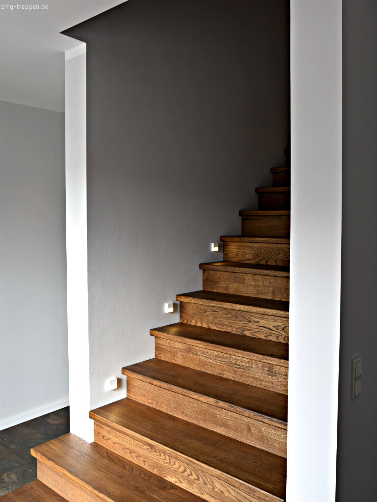 Moderne Holztreppe an grauer Wand mit kleinen quadratischen Wandlampen, führt nach oben.