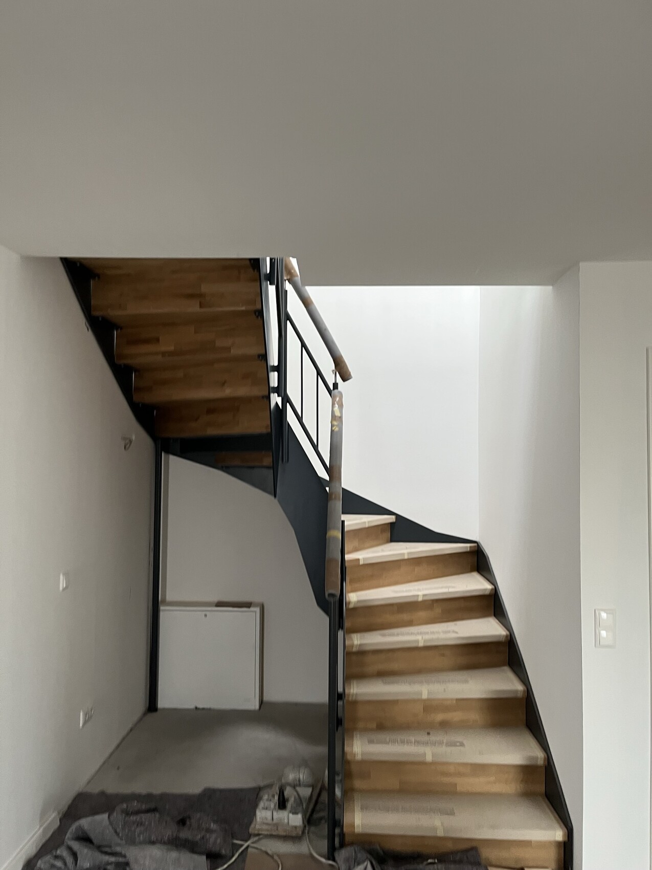 Treppenhaus unter Renovierung: Holzstufen, schwarzes Geländer, Baustellenmaterial am Boden.