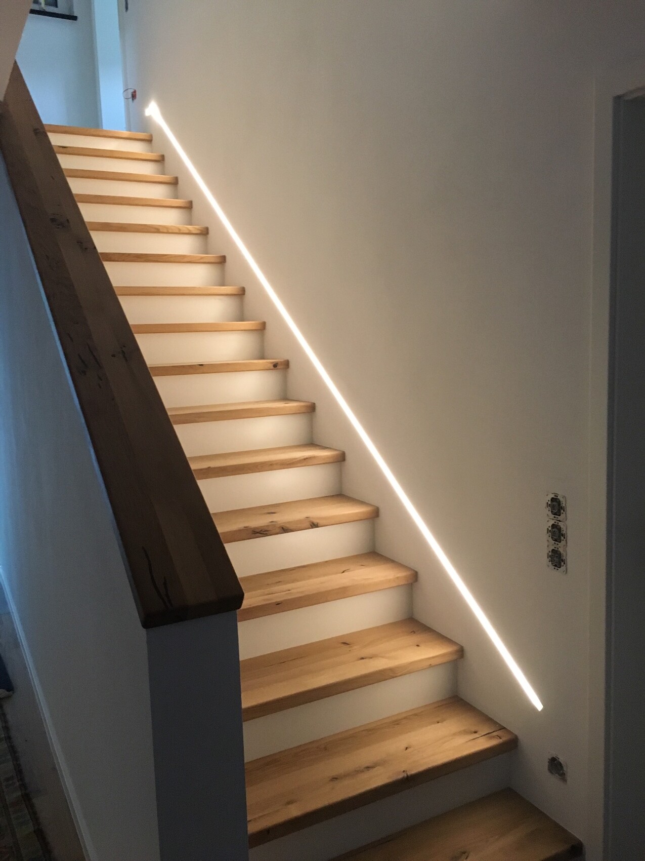 Moderne Holztreppe mit hellen Stufen, weißen Seitenwangen und LED-Lichtleiste an der Wand.