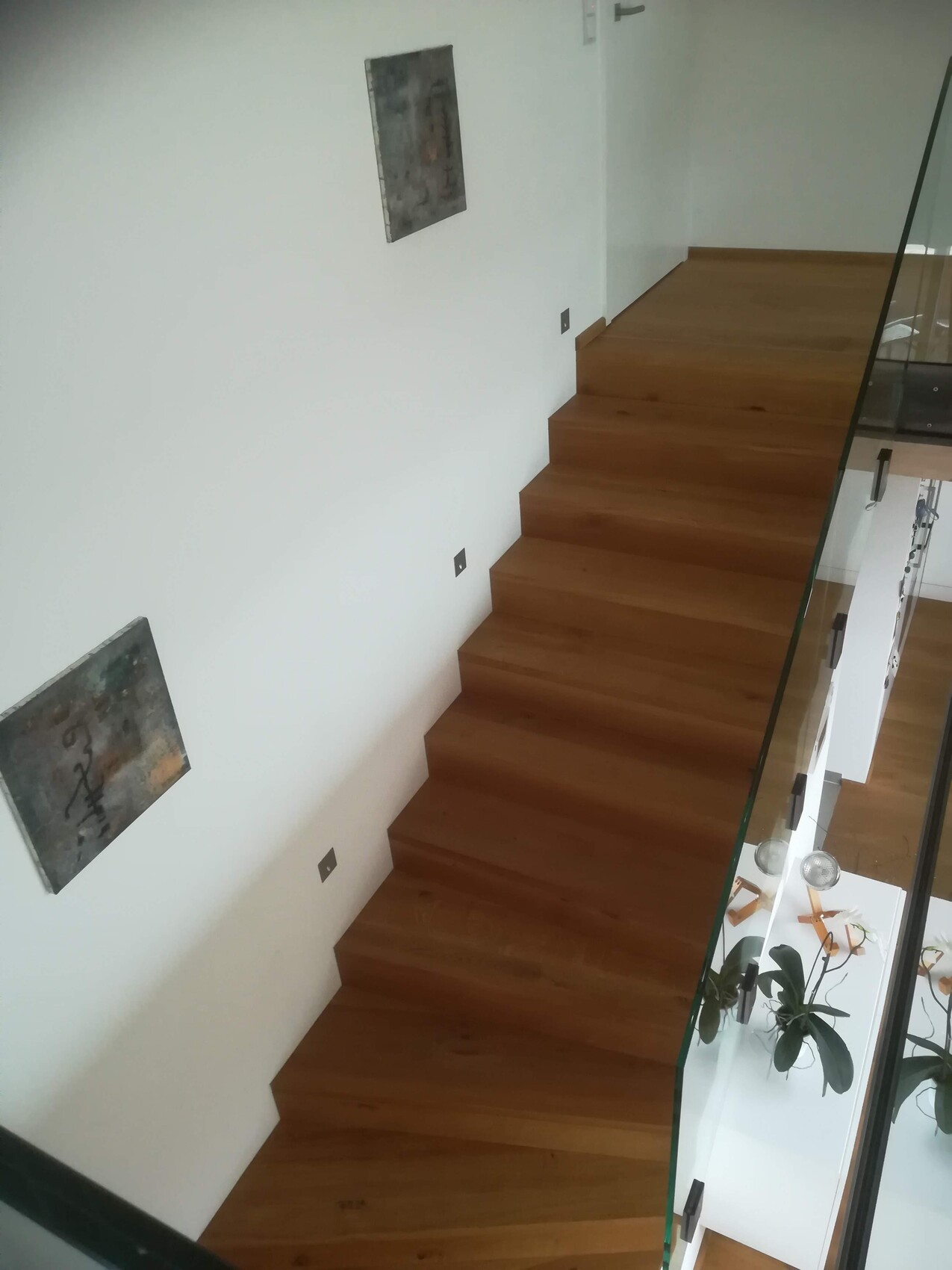 Moderne Holztreppe mit Glasgeländer an weißer Wand; quadratische Kunstwerke an der Wand.