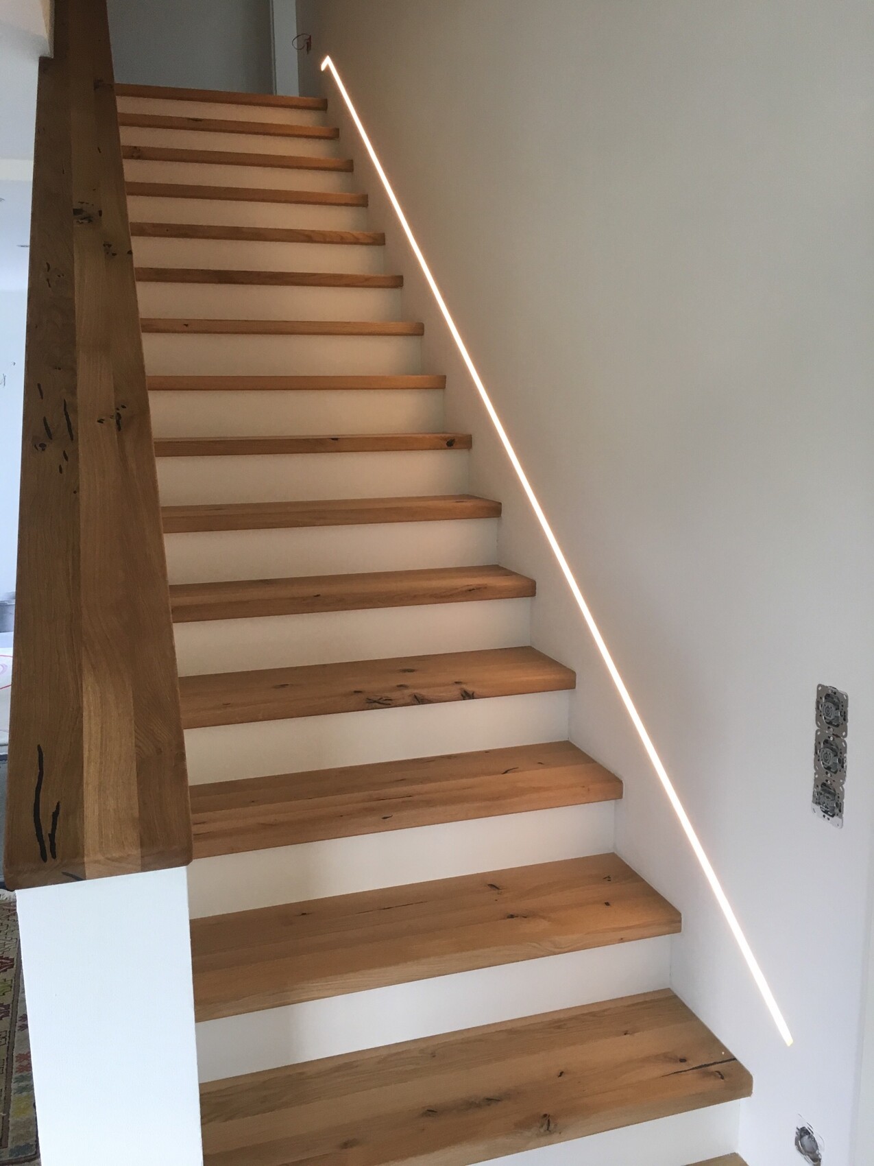 Moderne Holztreppe mit LED-Leiste an der rechten Wand, massiver Holzpfosten links, weiße Wände.