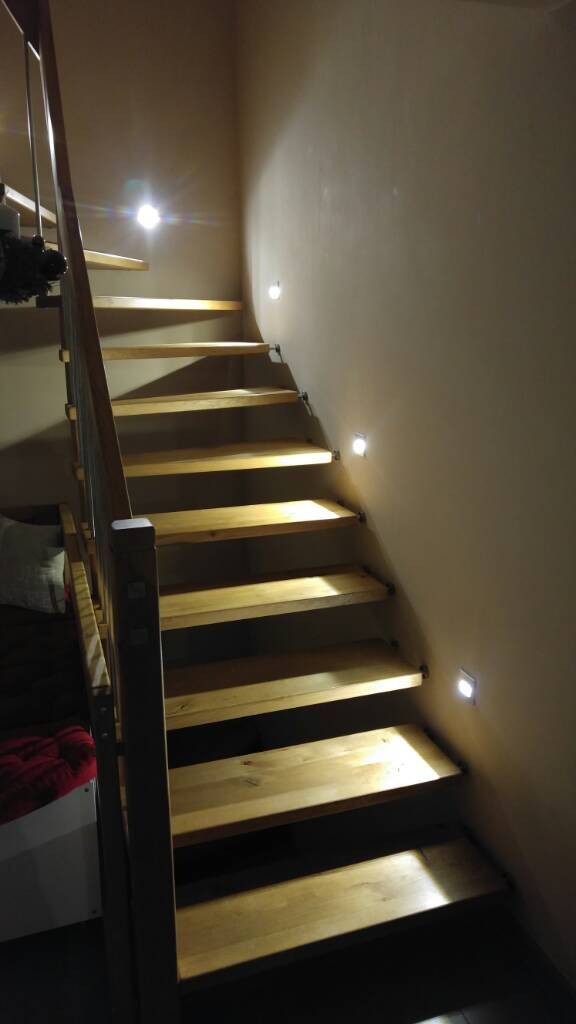 Holztreppe mit offenen Stufen, seitliche Wandlichter beleuchten die Treppe.