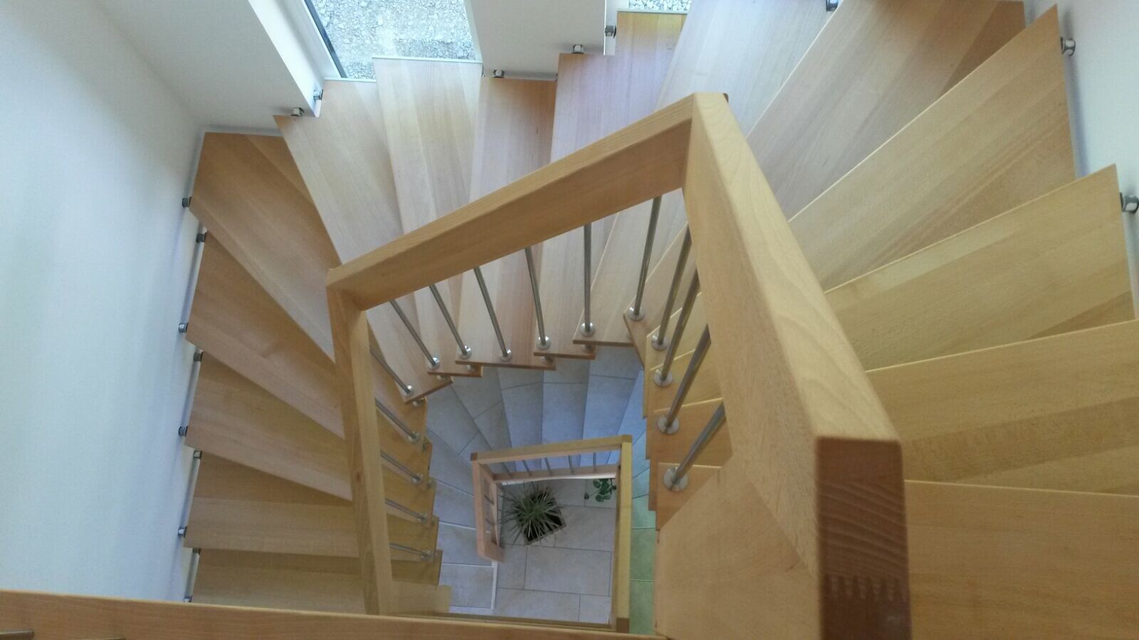 Obere Sicht auf eine helle Holztreppe mit Metallstab-Geländer, Blick nach unten auf eine Pflanze am Boden.