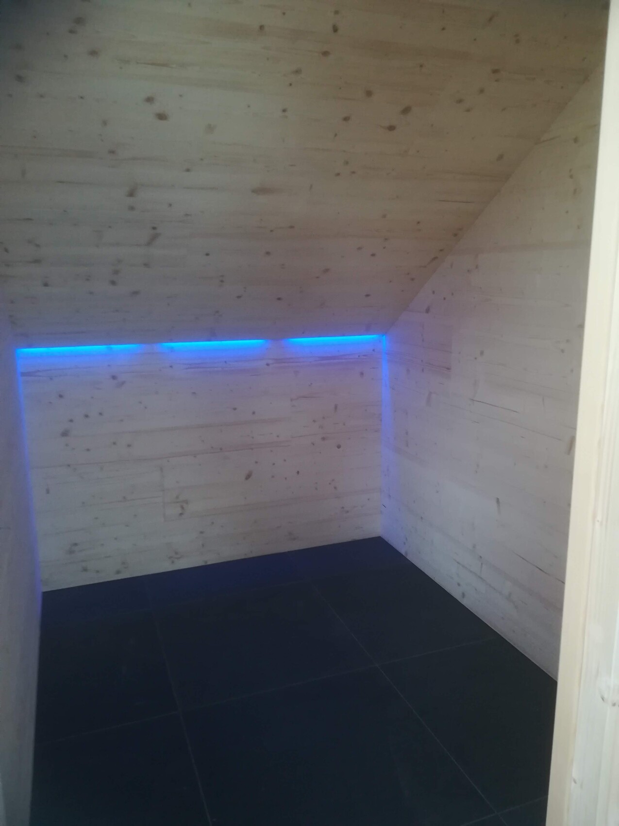 Kleiner Raum mit Holzverkleidung, blaue LED-Lichtleiste entlang der Ecke, dunkler Boden.