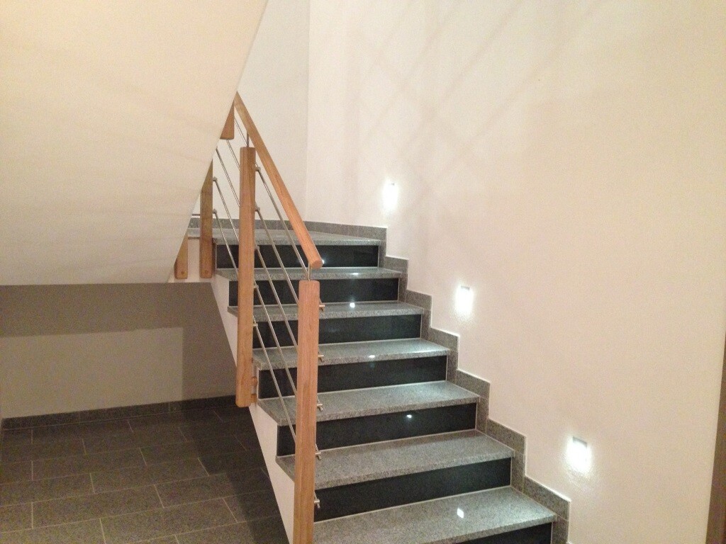 Innenraum: Treppe mit grauen Stufen, Holzhandlauf, helle Wände und Wandlichter.