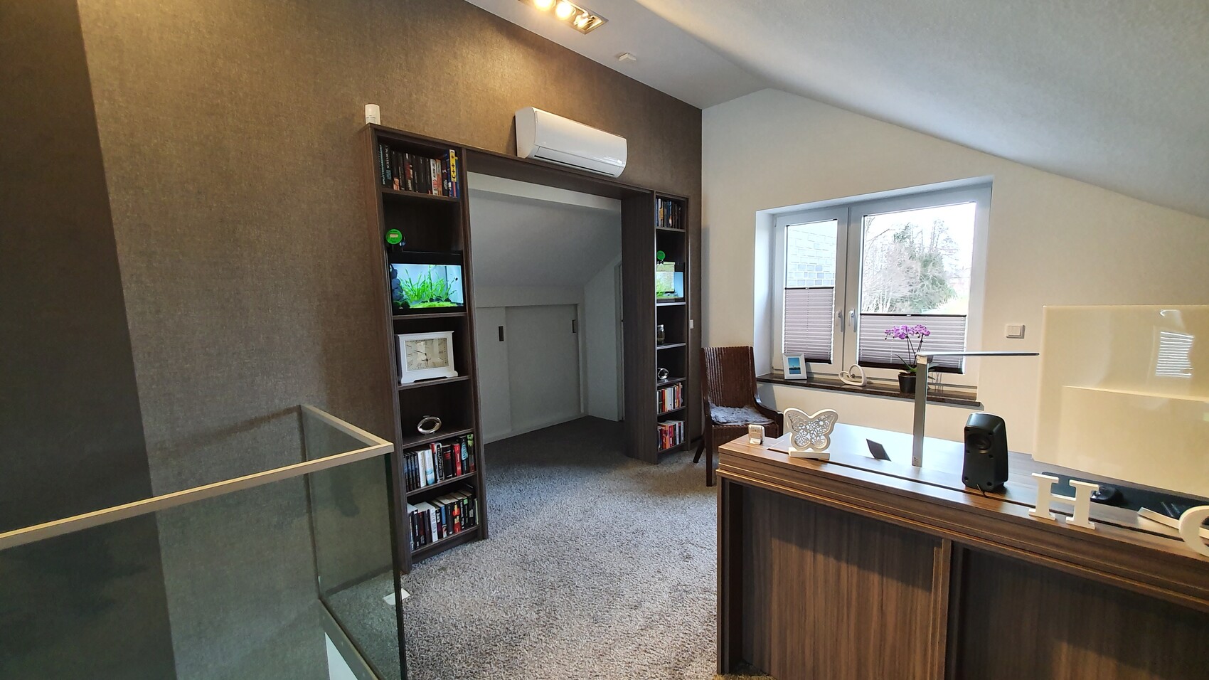 Wohnzimmer mit dunkler Regalwand, Durchgang, Aquarium, Teppichboden, Fenster mit Orchidee.