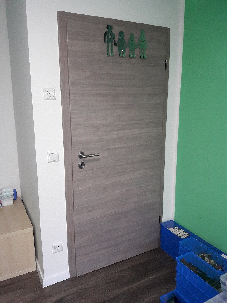 Holztür mit grünem Familien-Symbol-Aufkleber oben, weiße Wand links, blaue Kisten unten.