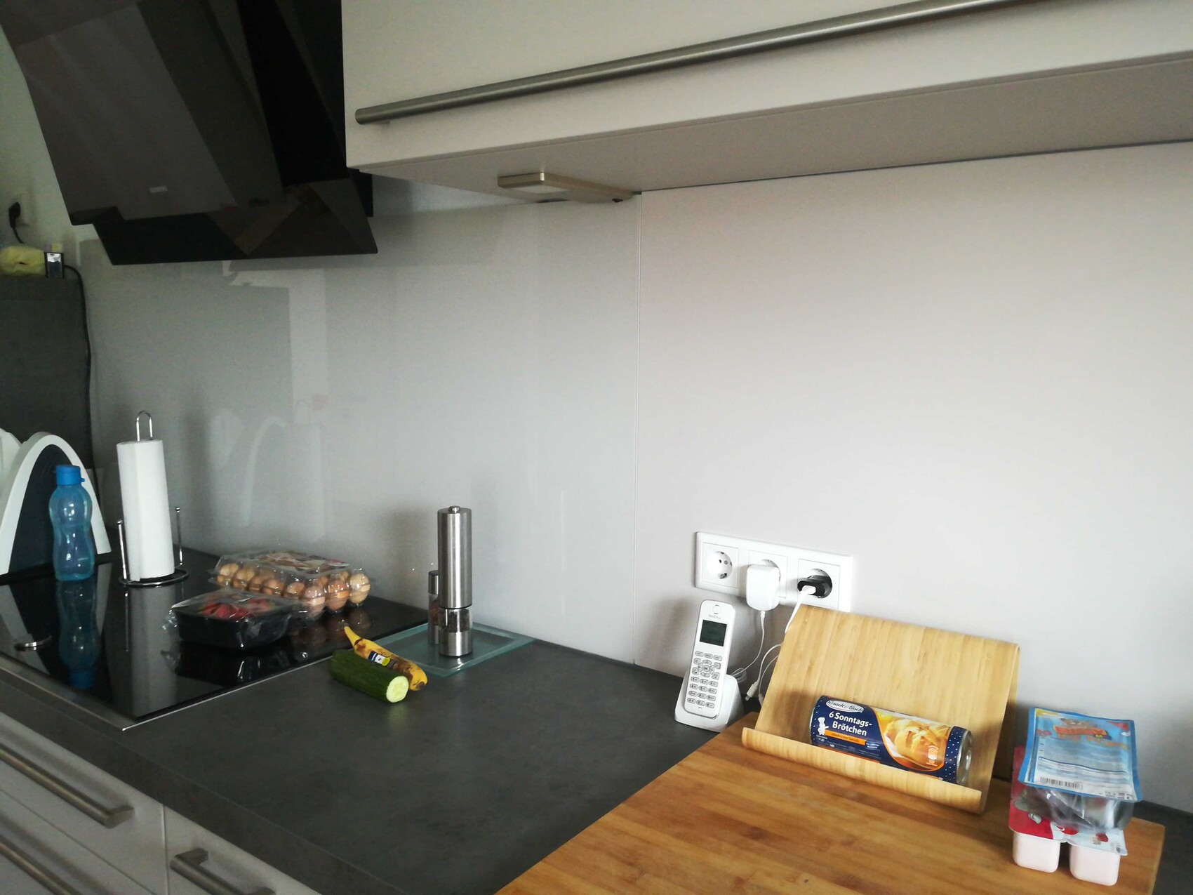 Moderne Küchenzeile mit Glasrückwand, Arbeitsplatte, Eierkarton, Gurke, Küchenrolle und Telefon.