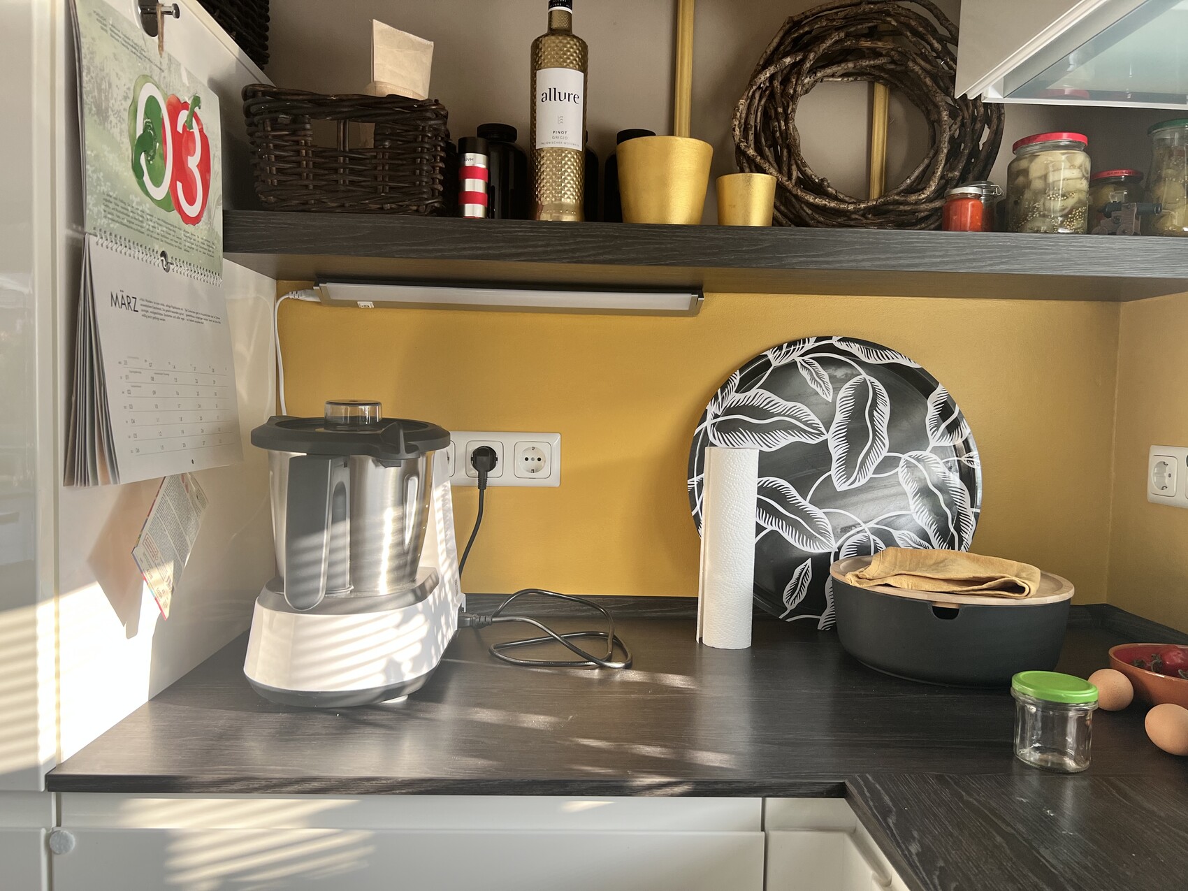 Küche: Entsafter links, Papierrolle, Topf, schwarze Platte mit weißen Blattmotiv vor gelber Wand.