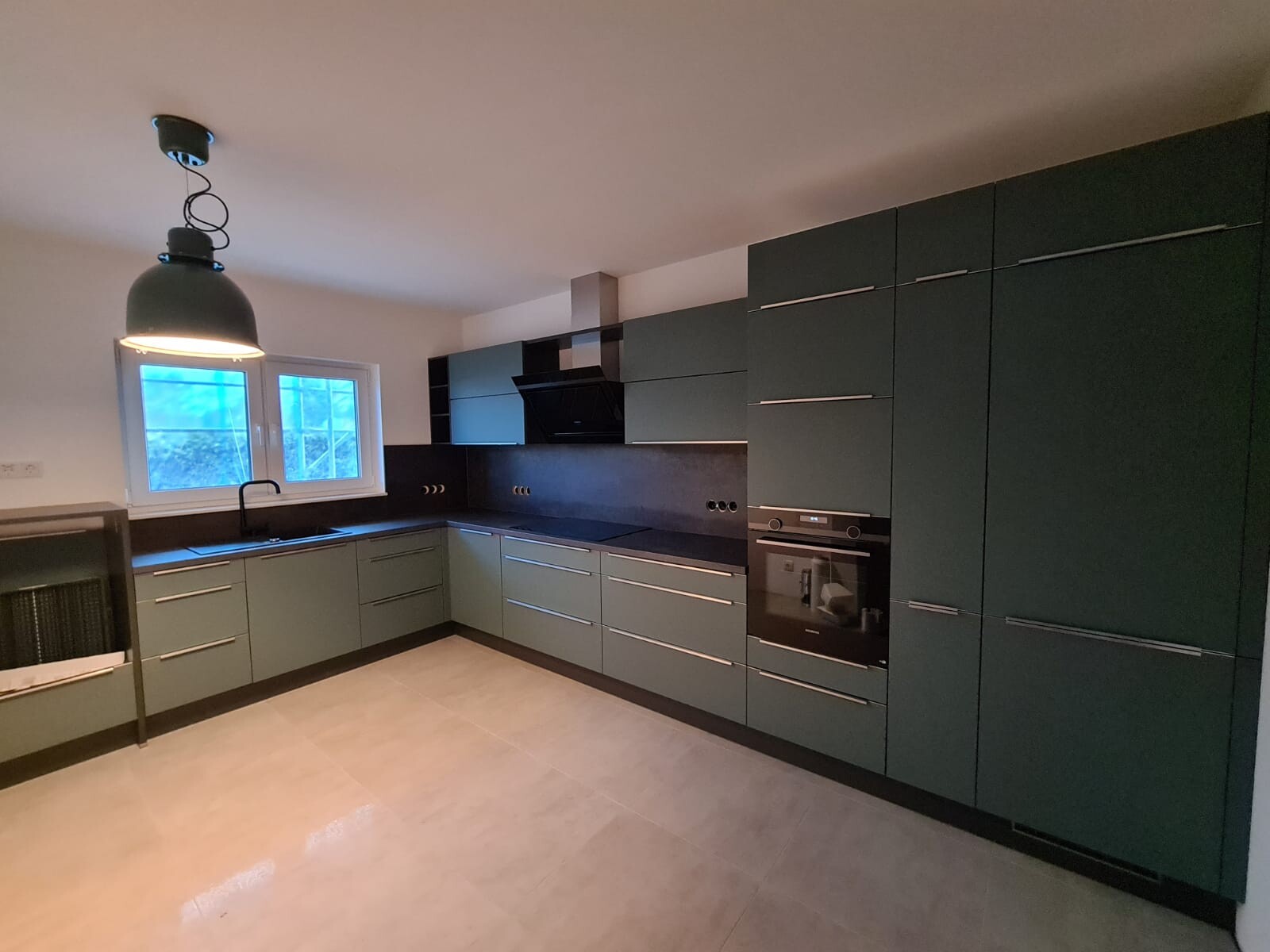 Moderne Küche mit grünen Fronten, schwarzer Arbeitsplatte, Backofen, Dunstabzug und Spüle am Fenster.