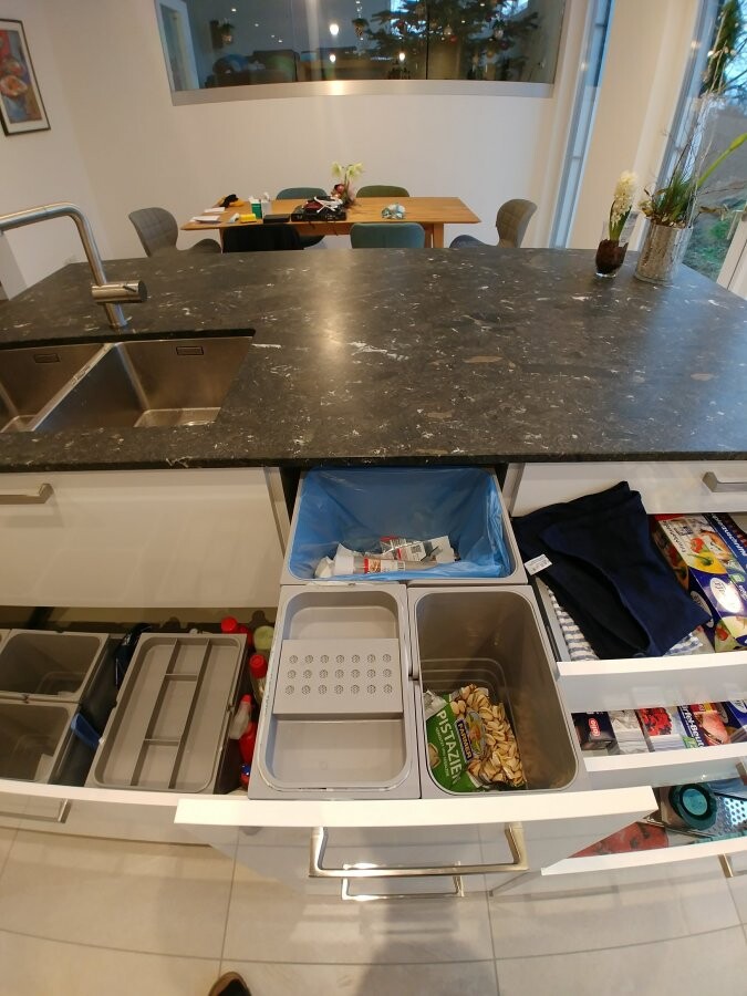 Küche mit dunkler Arbeitsplatte, Spüle, offene Schubladen und Müllbehälter; Esstisch im Hintergrund.