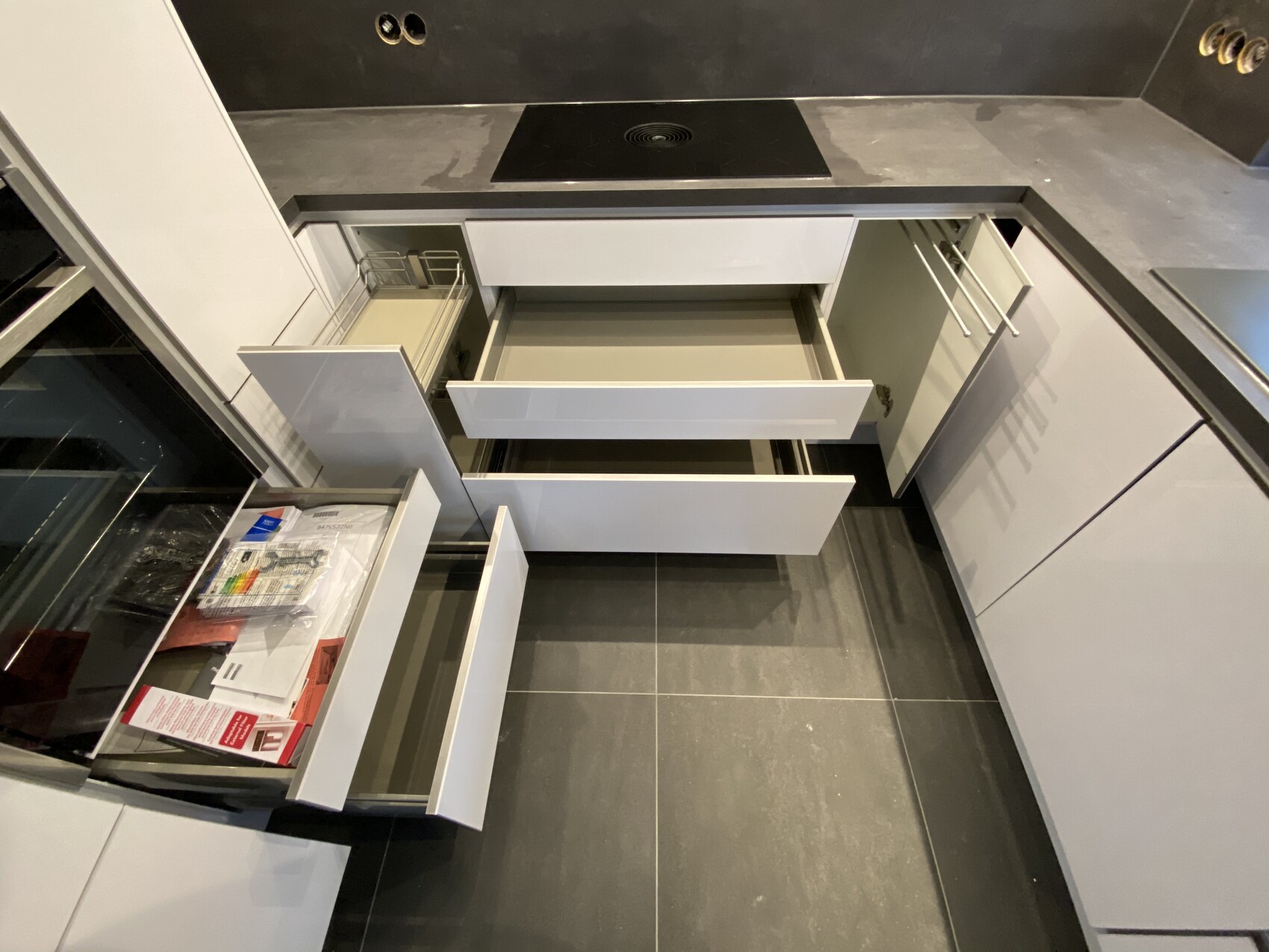 Moderne Küche mit dunkler Arbeitsplatte, Induktionskochfeld und geöffneten weißen Unterschränken.