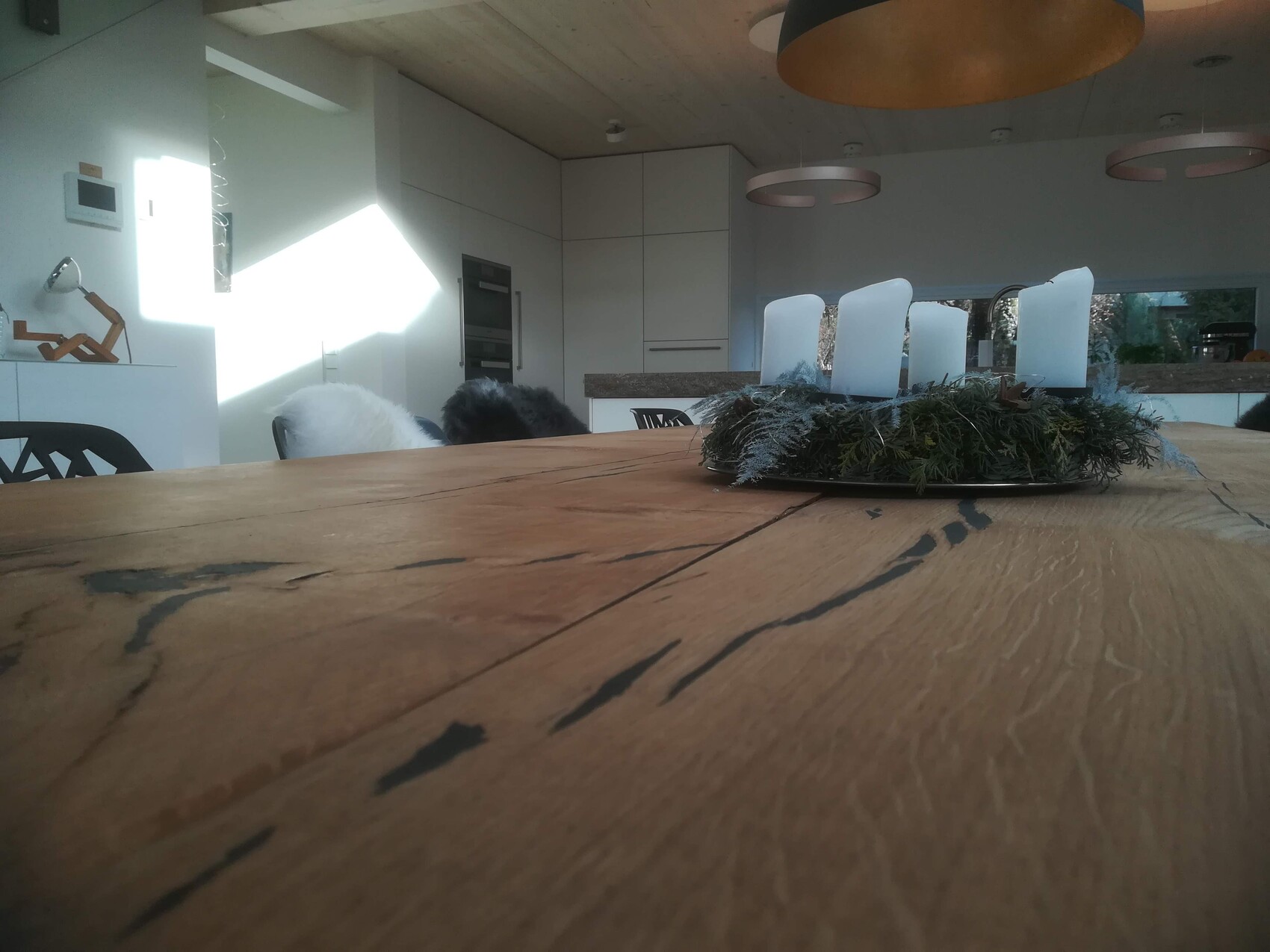 Holztisch mit grünem Kranz und vier weißen Kerzen, moderne Küche im Hintergrund.