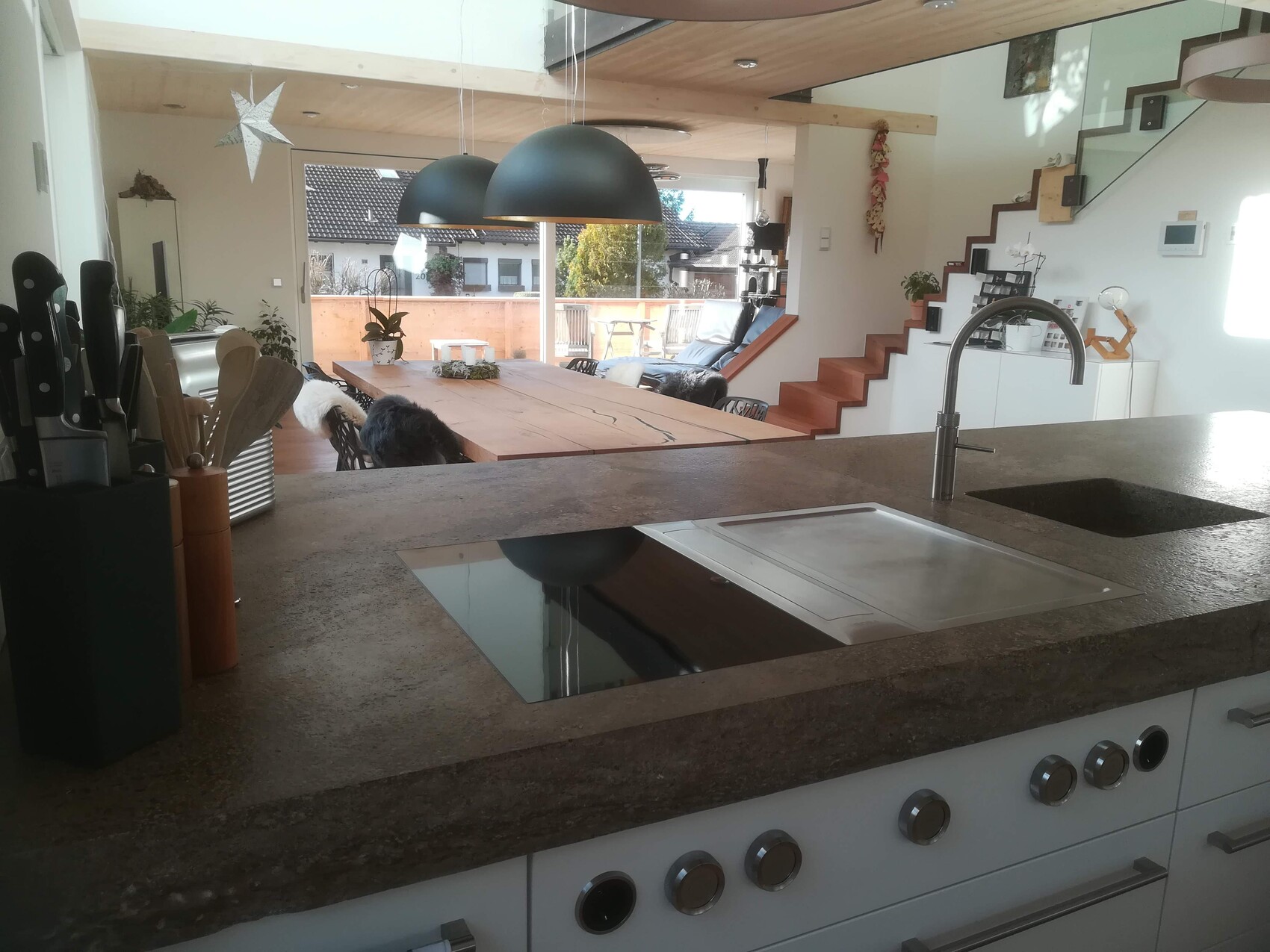 Offene Küche mit Insel, Spüle, Induktionskochfeld, großer Holztisch, Pendelleuchten, Treppe.