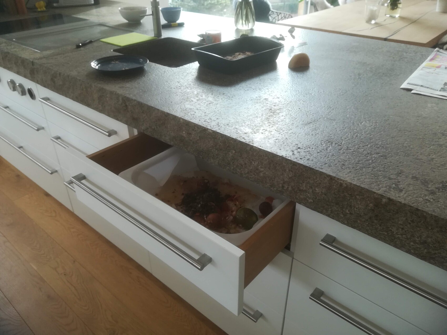 Moderne Kücheninsel mit grauer Granitplatte; offene Schublade zeigt Essen/Behälter, daneben Teller.