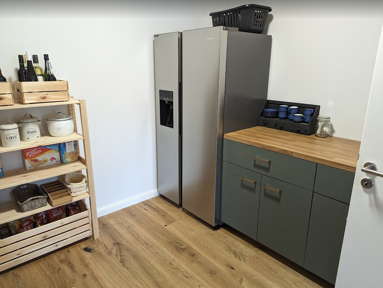Küche: großer Edelstahl-Kühlschrank, Holzregal mit Flaschen, grüne Schränke, blauer Bechersatz.