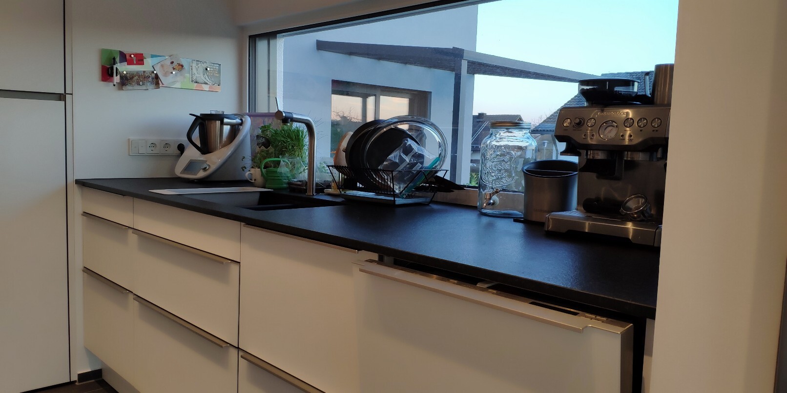 Moderne Küche mit Spüle, Geschirrkorb, Kaffeemaschine, Gläser, Pflanze vor dem Fenster.