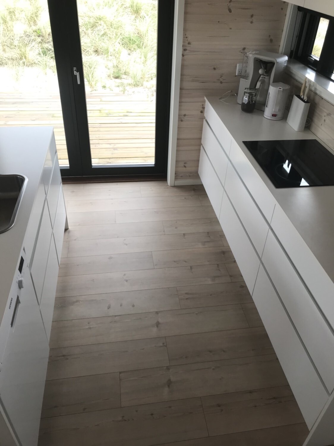 Moderne weiße Küche mit Induktionskochfeld, Spüle, Geräten, Holzboden und Glastür nach draußen.