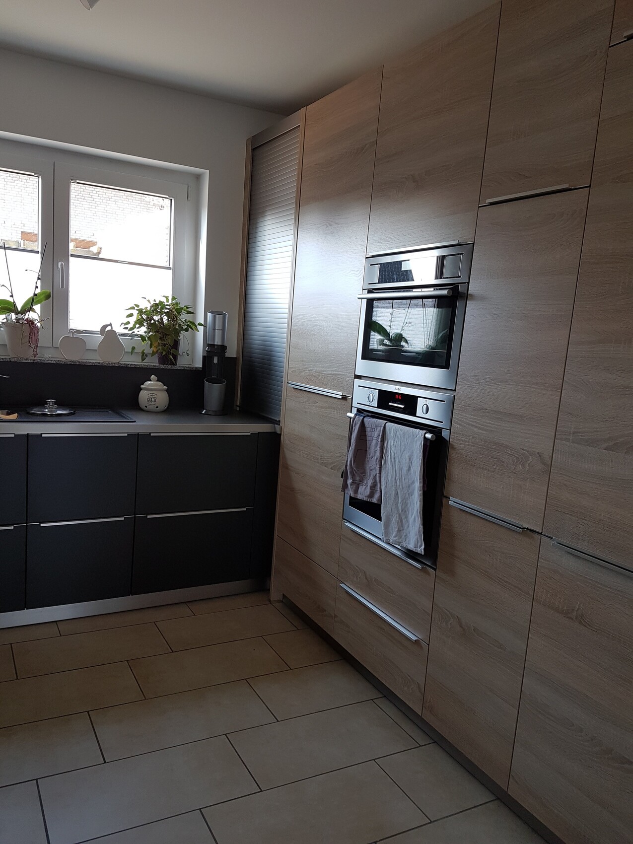 Moderne Küche: eingebauter Backofen, Holzfronten, dunkle Unterschränke, Fensterbank mit Pflanzen.]