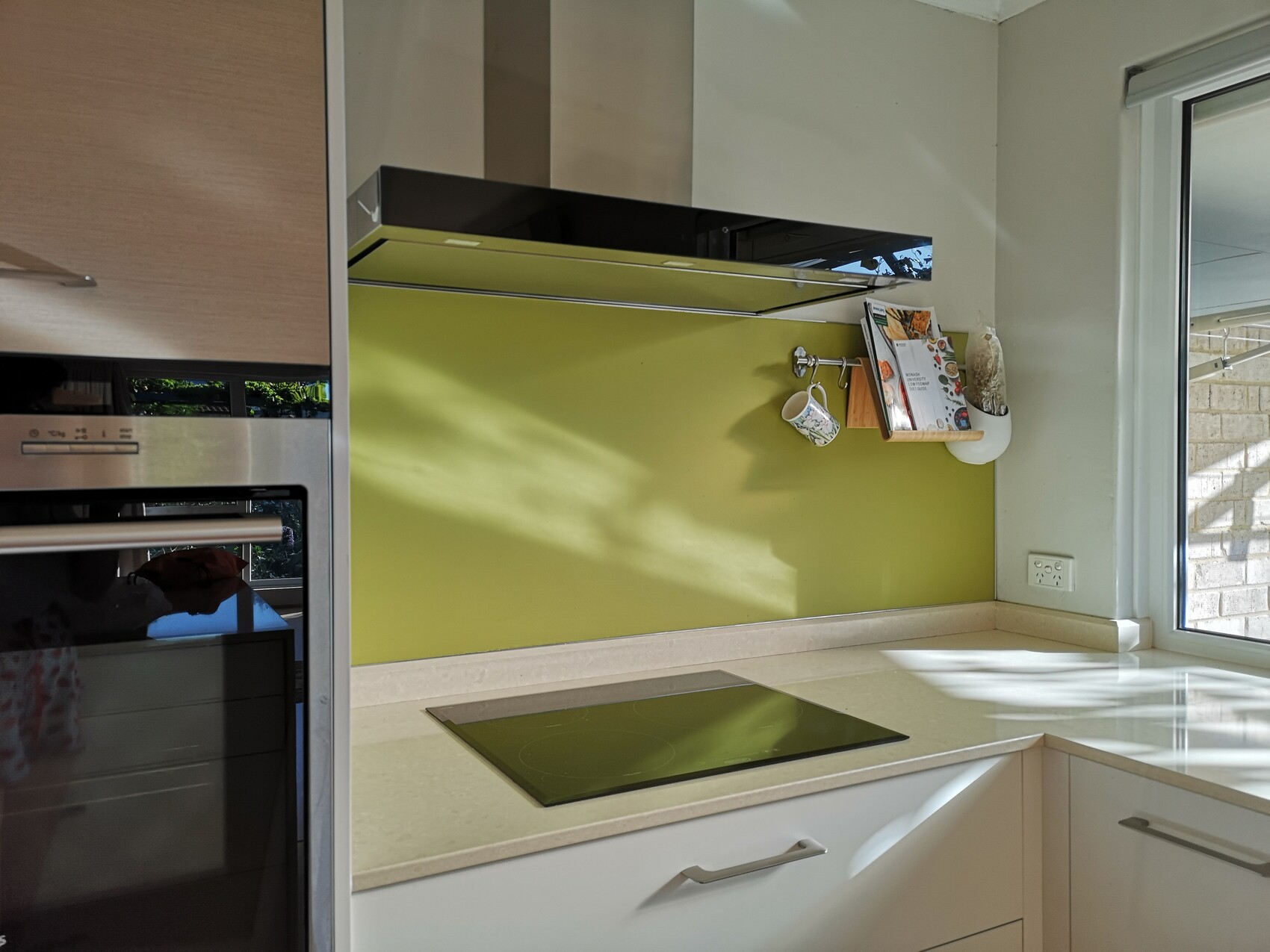 Moderne Küche mit grüner Glasrückwand, Induktionskochfeld und Wandregal mit Kochbuch und Tasse.