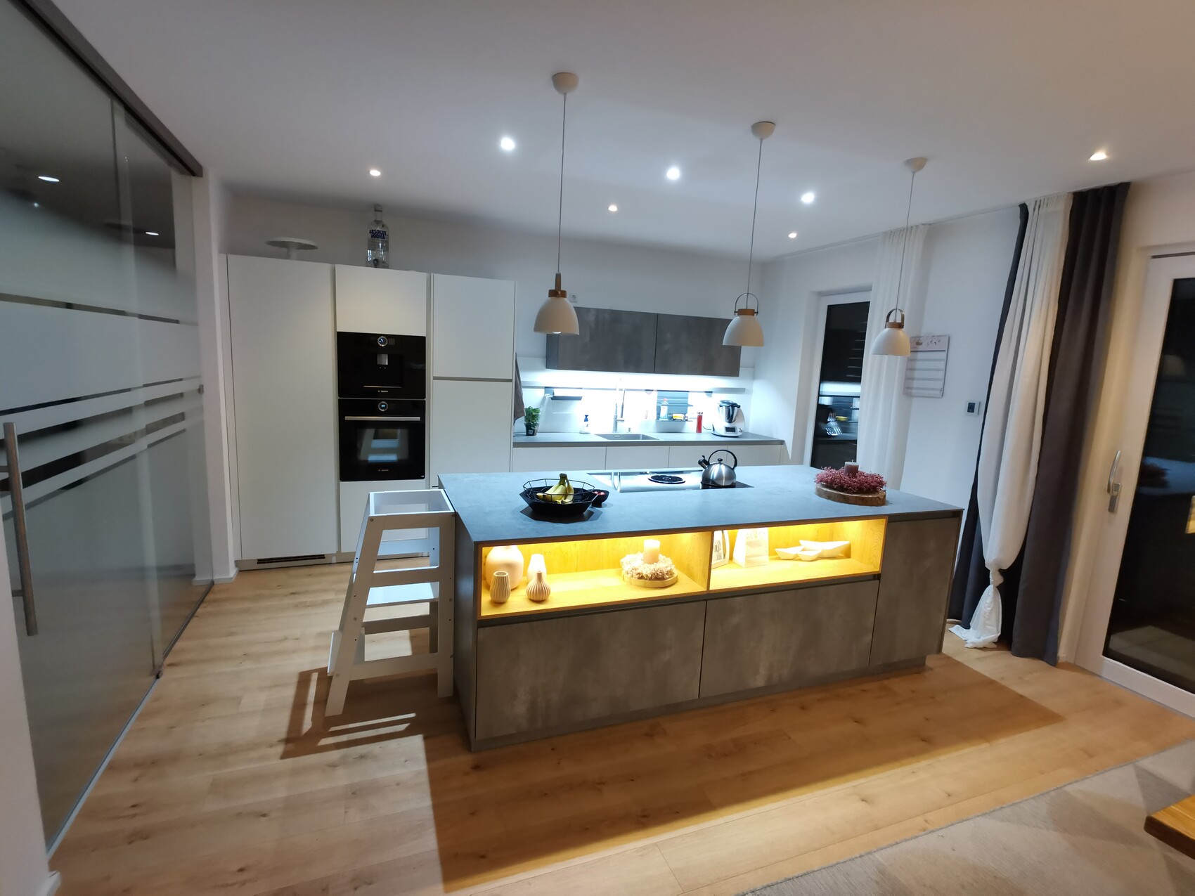 Moderne Küche mit zentraler Insel, Pendelleuchten, Backofen, weißen Schränken und Holzboden