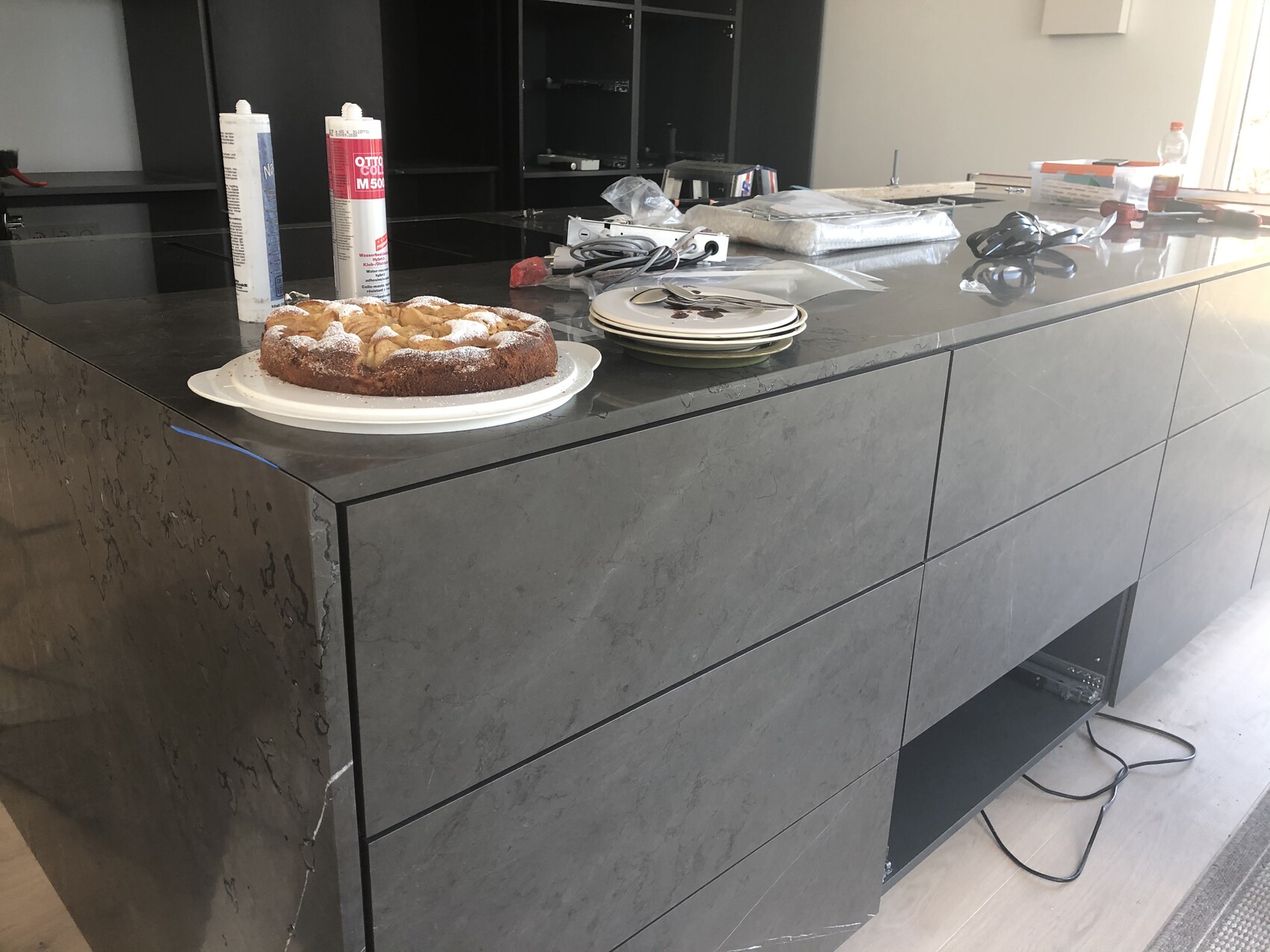 Kuchen auf dunkler Kücheninsel, daneben Kleberdosen, Tellerstapel und Kabel im Hintergrund